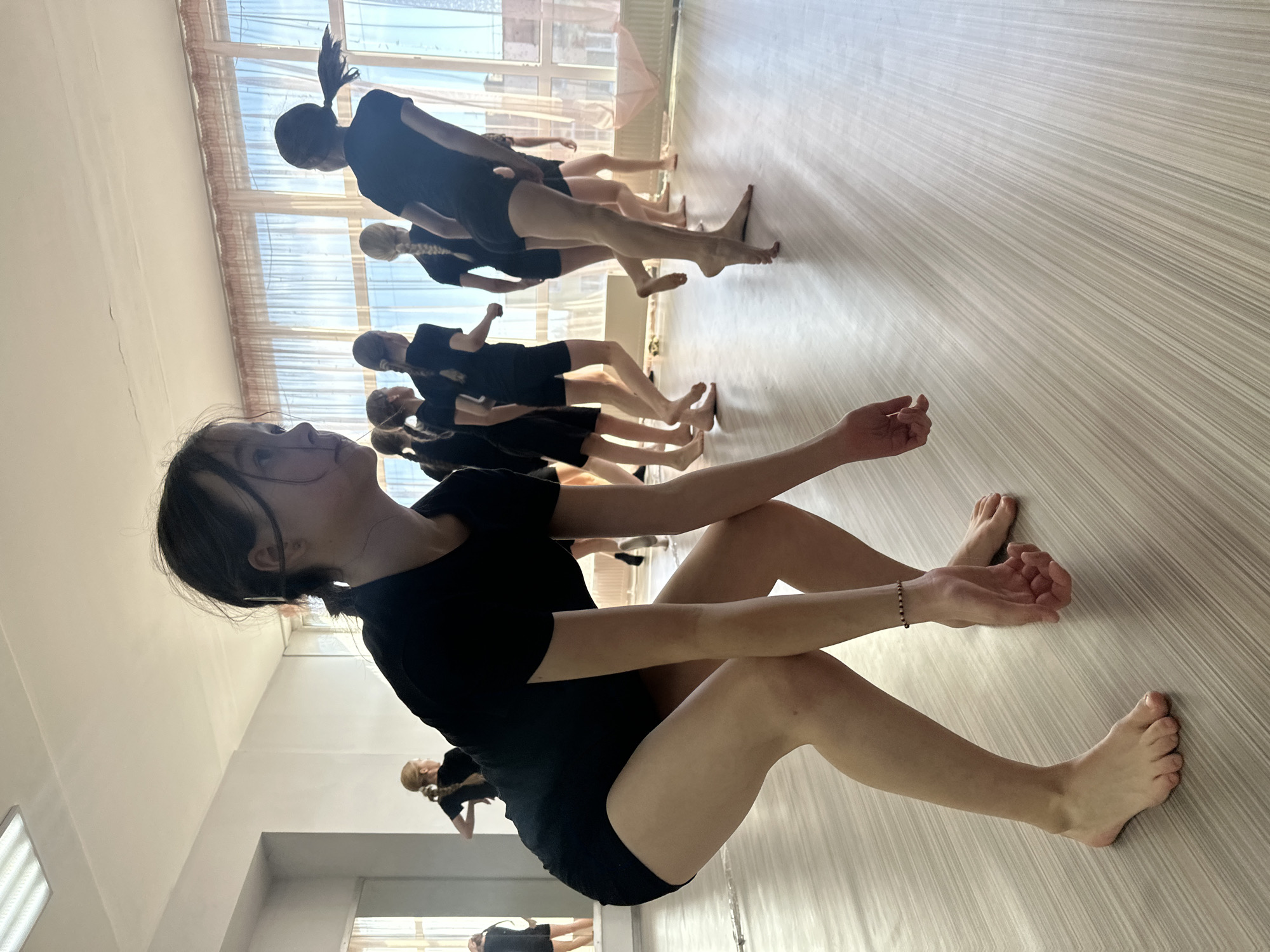 A medium shot depicts a dance class in Votkinsk, Russia. The class is taking place indoors. A group of people are participating in the class, all wearing black dance attire. The camera angle is positioned low. The focal point is a person seated in the foreground. Other participants are standing in the background performing a dance routine. The room has a patterned floor, and there is a window.