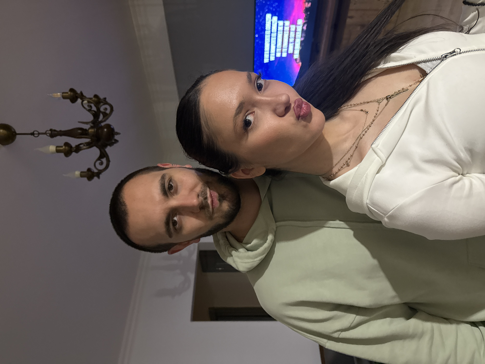 A couple is posing for a photo indoors. The man has short dark hair and a beard, and is wearing a light green hoodie. The woman has long dark hair, and is wearing a white zip-up top with necklaces. She is making a kissing face. A chandelier is visible in the background to the left. A TV screen showing colorful patterns is visible in the background above the woman. The image appears to be taken in Yerevan, Armenia.