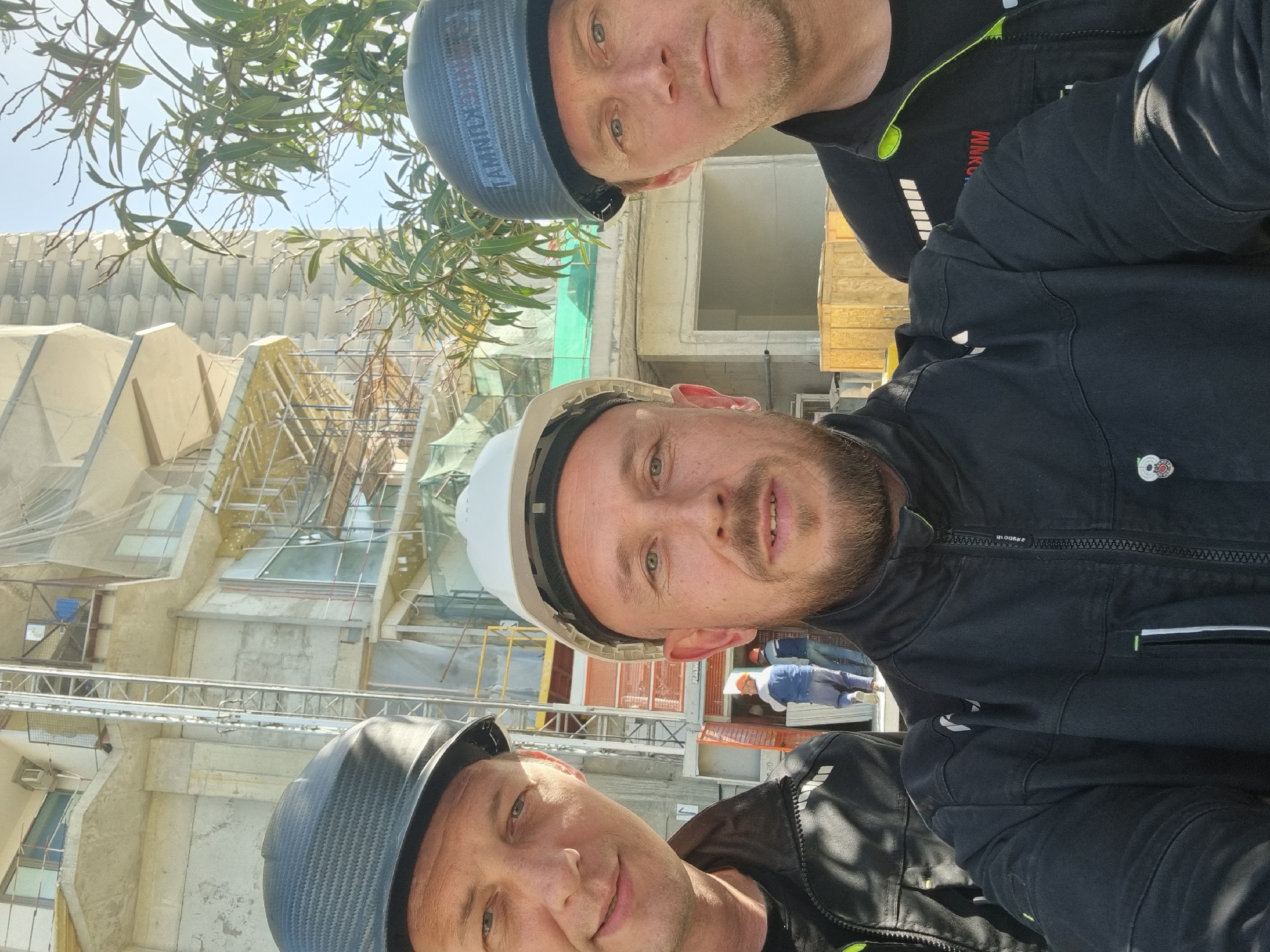 Three construction workers are taking a selfie at a construction site in Sochi, Russia. The workers are wearing hard hats and work clothes. The building under construction has scaffolding around it. Other workers can be seen in the background.