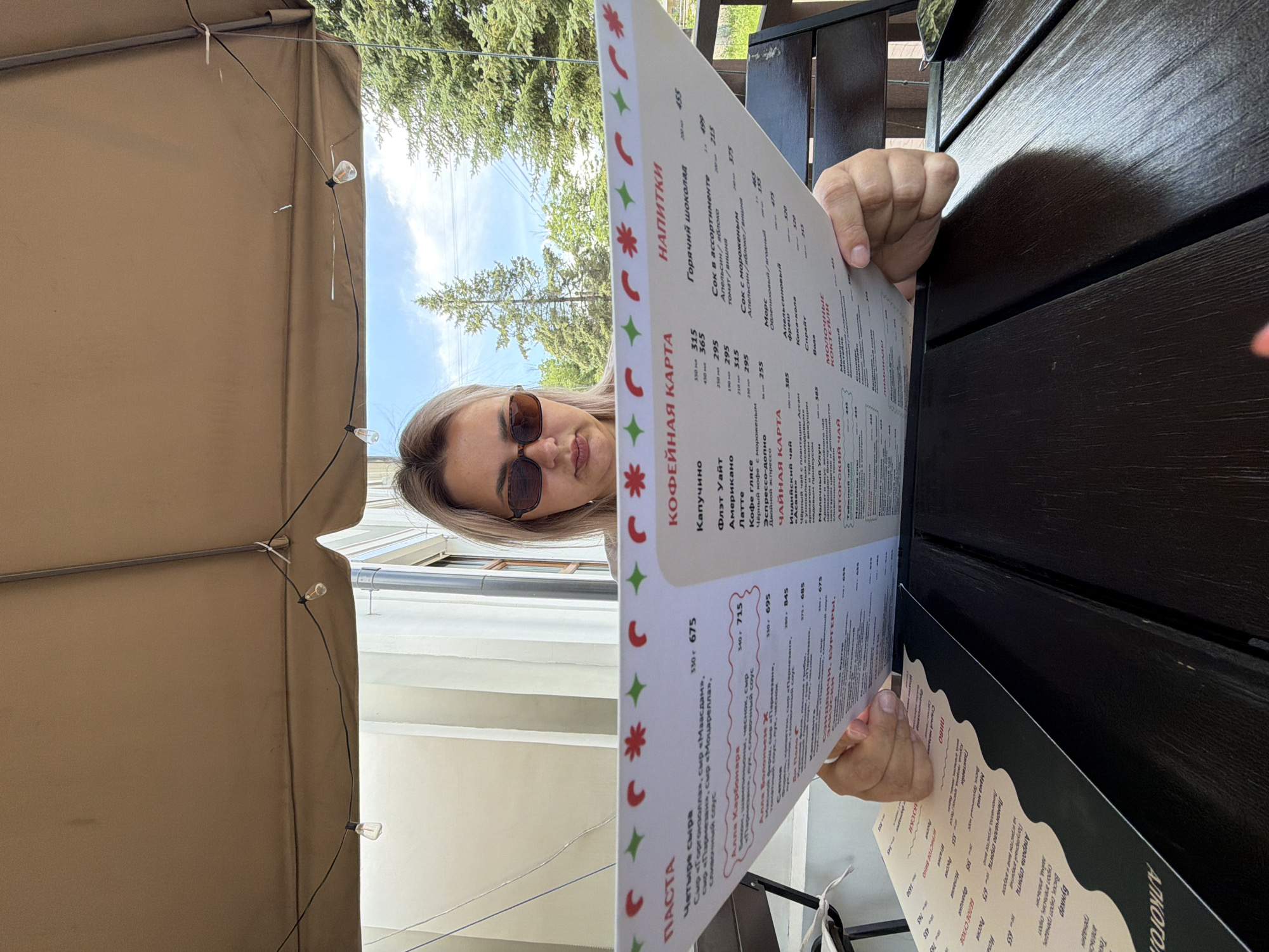 A young woman with blonde hair and sunglasses is sitting at an outdoor table and reading a menu. She is positioned in the foreground, with the menu obscuring much of her body. The menu is large and appears to be made of laminated paper, with various food and drink items listed in Russian. Behind the woman, a wooden umbrella provides shade, and string lights are visible. Beyond the umbrella, trees and a building are visible under a blue sky. The scene appears to be at a cafe or restaurant. There is no specific information in the image to indicate the location is Birobidzhan, Russia.