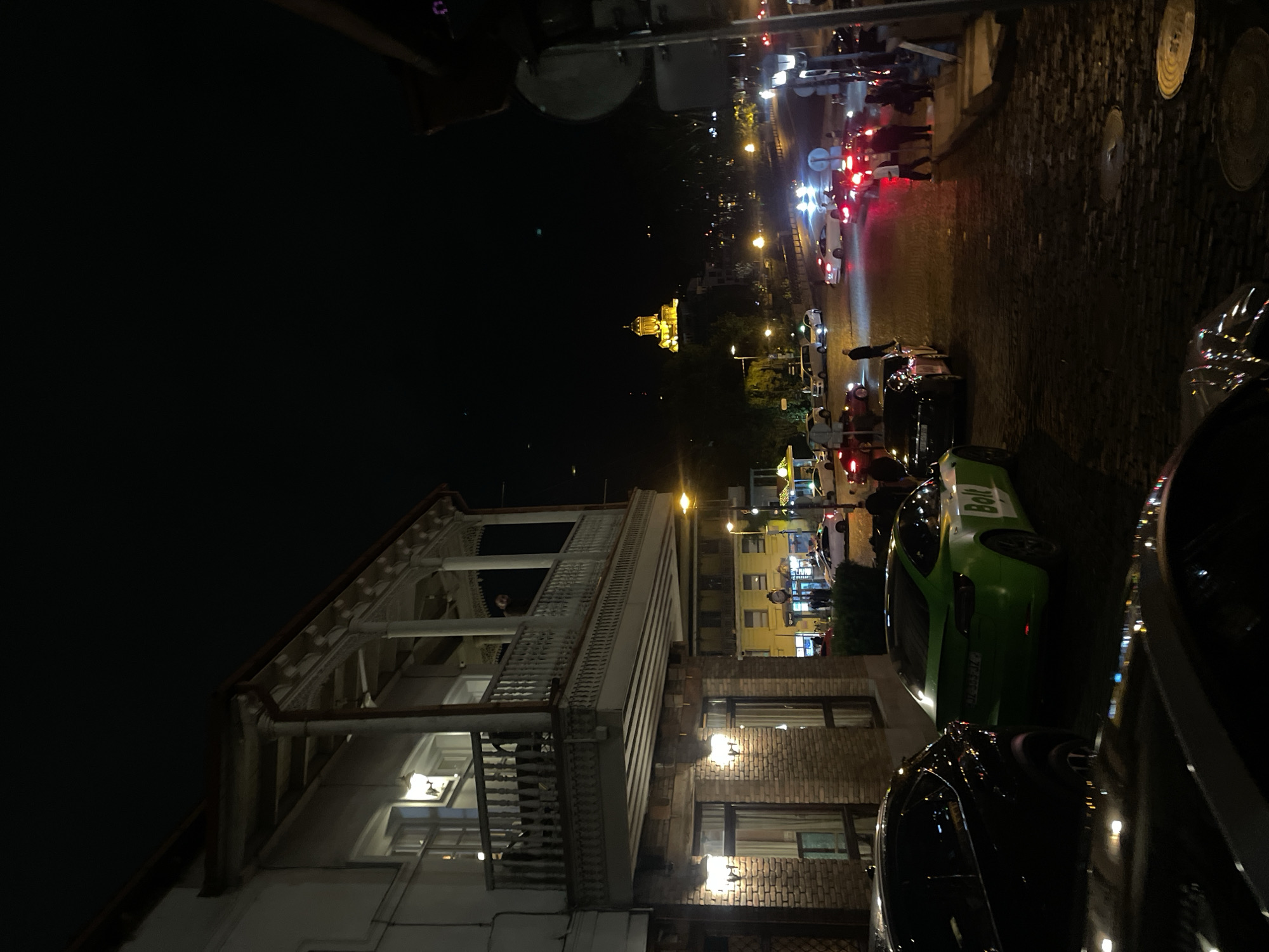 The image captures a night scene in Tbilisi, Georgia. It shows a building with a traditional Georgian balcony, lit by interior and exterior lights. Adjacent to the building, a street scene unfolds with several cars parked along the side, one of which is a green "Bolt" vehicle. In the distance, the illuminated Holy Trinity Cathedral of Tbilisi is visible against the dark sky. People are observed crossing the street at a crosswalk. The street appears to be paved with cobblestones. The lighting is a combination of streetlights, car headlights, and building illumination.