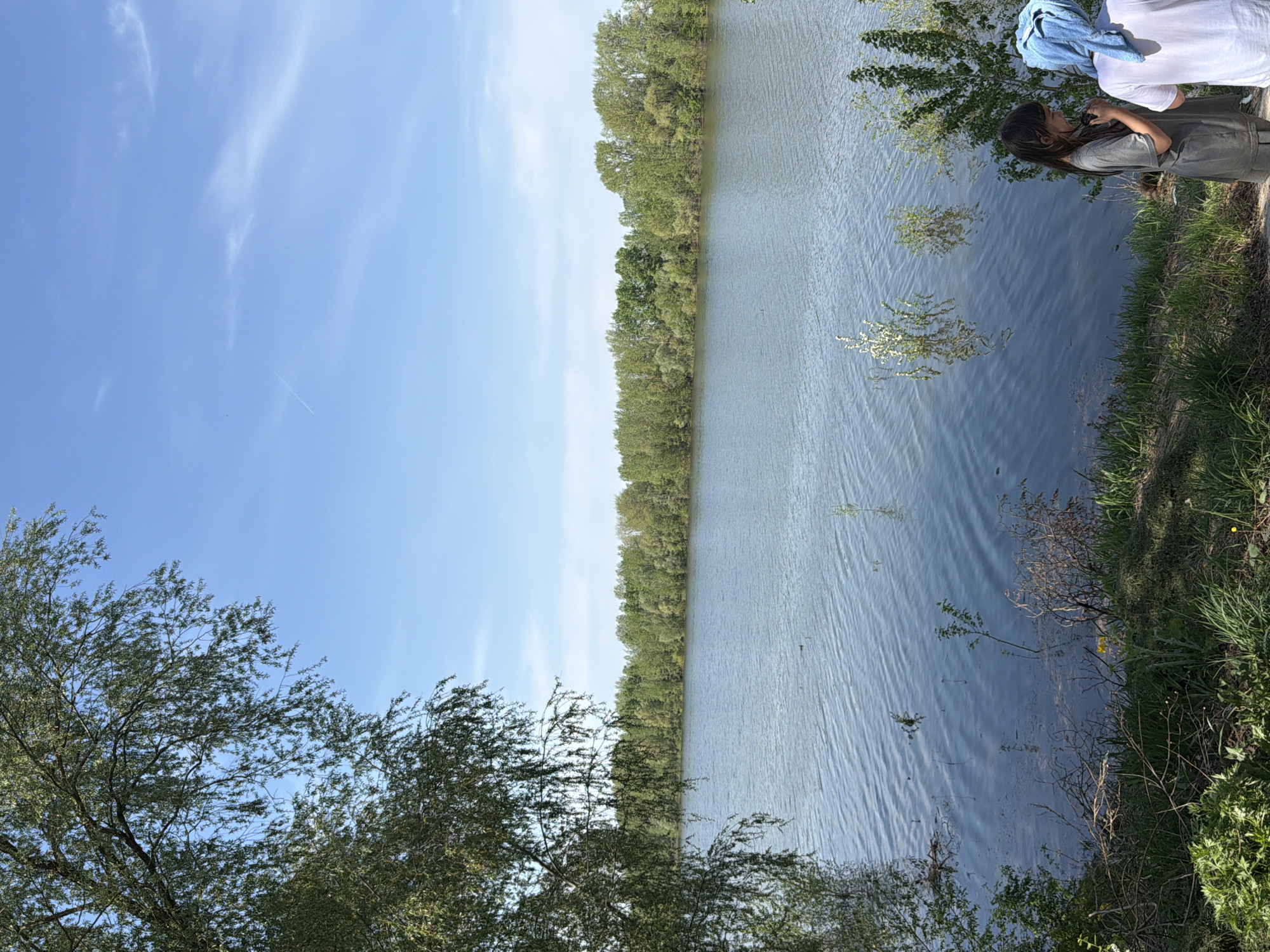 The image shows a body of water with trees lining the opposite bank. The sky is blue with some clouds. In the foreground, there are trees and vegetation. A person stands near the water's edge, possibly taking a picture. The location is identified as the city of Chaikovsky, Russia.