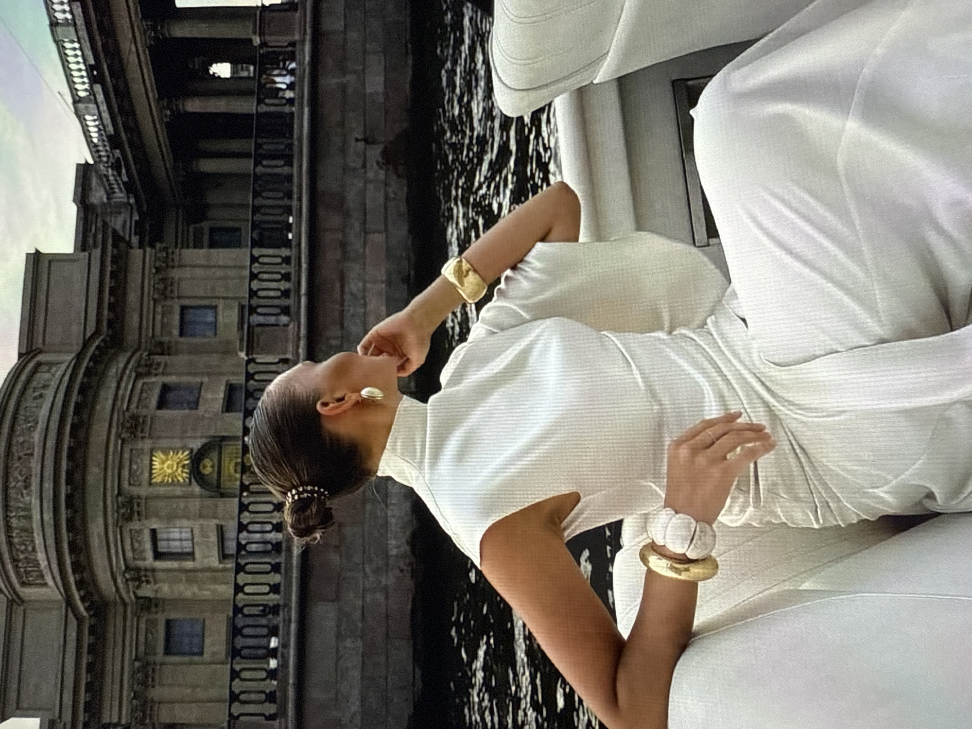 A woman with her hair in a bun is seated on a white upholstered seat, possibly a boat, with her back to the camera. She is wearing a white outfit, including a high-necked top with short sleeves and wide sleeves, and a long skirt. She has a gold oval earring in her visible ear and two bracelets on her right wrist, one gold and one white. Her right hand rests on her lap, and her left hand is bent at the elbow, with her fingers near her face. In the background, a large, ornate stone building with columns and a balustrade is visible across a body of water. The sky above is overcast. There is no identifiable indication of the city of Găvănoasa, Moldova in the image.