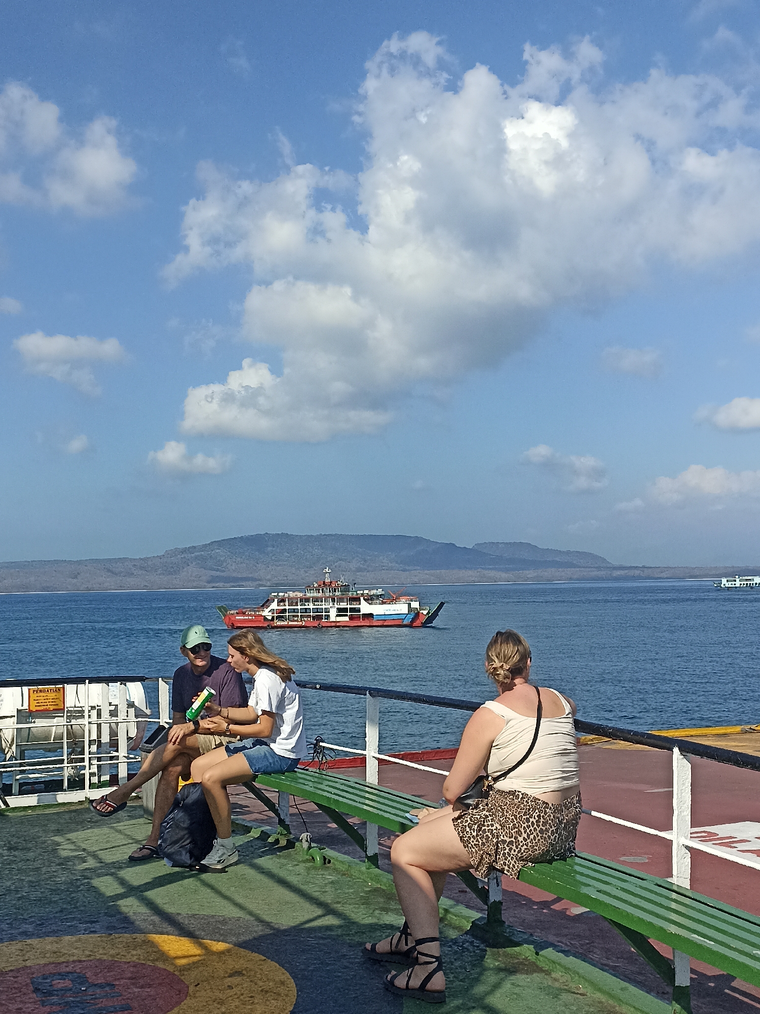 On a sunny day with scattered clouds, two people sit on a bench on a ferry deck. A man wearing a purple t-shirt, green cap, and sunglasses sits to the left, with a backpack at his feet. A young woman with blonde hair, wearing a white t-shirt and denim shorts, sits to his right. They appear to be talking, with the woman holding a green bottle. In the background, a large red and white ferry sails on the blue ocean, with a hilly, tree-covered island in the distance. Another smaller white boat is visible further out at sea. The ferry deck is wet and reflective, with markings on the floor. This scene is likely taking place in the waters near Ketapang, Indonesia, a port city known for its ferry services.