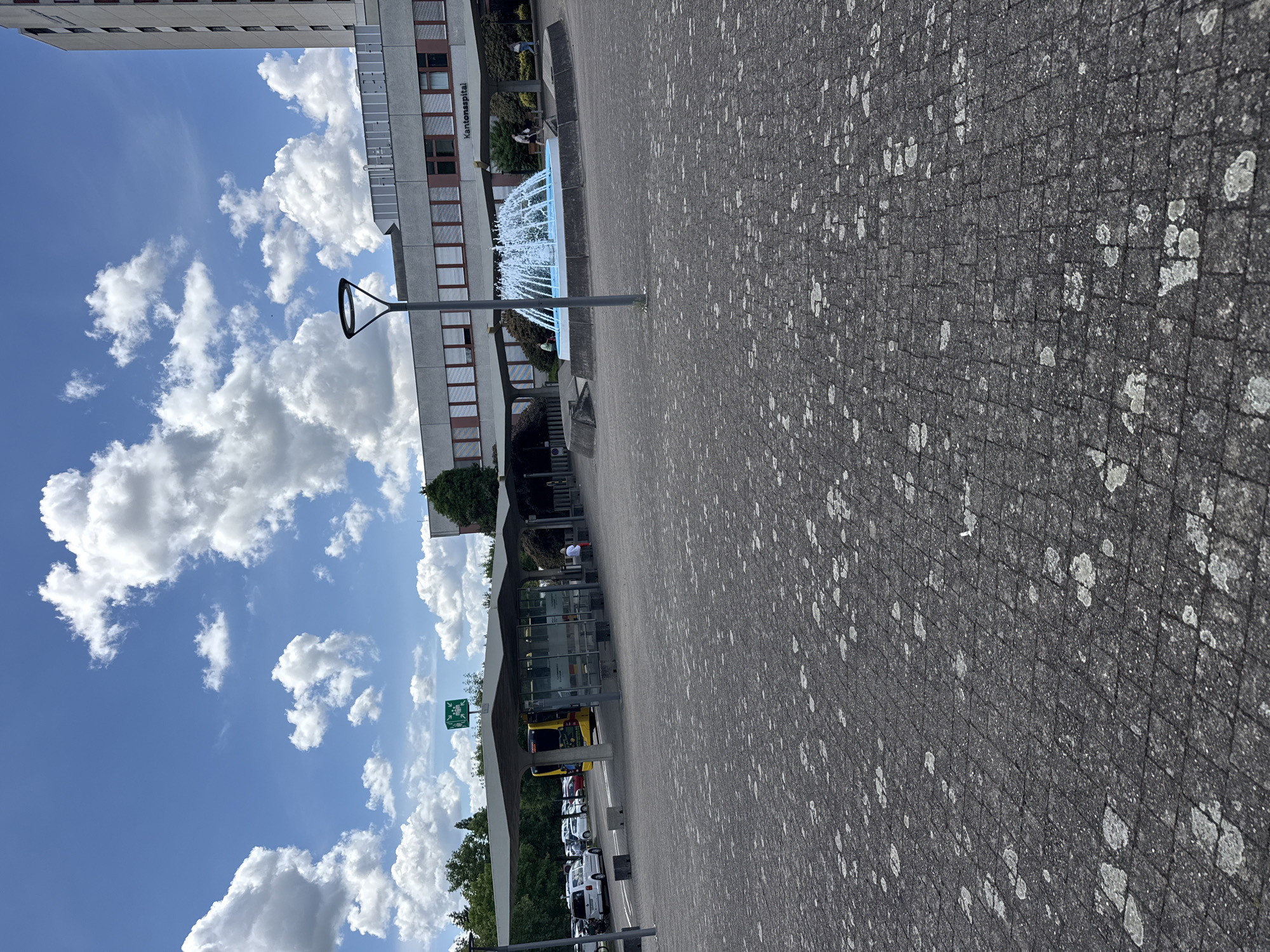 The image shows a street scene in Bottmingen, Switzerland, in front of the Kantonsspital. A fountain is visible. A bus stop is located in the foreground with a bus. Several cars are parked nearby. The sky is blue with cumulus clouds. The pavement is grey with white markings. There are some people nearby at the bus stop.