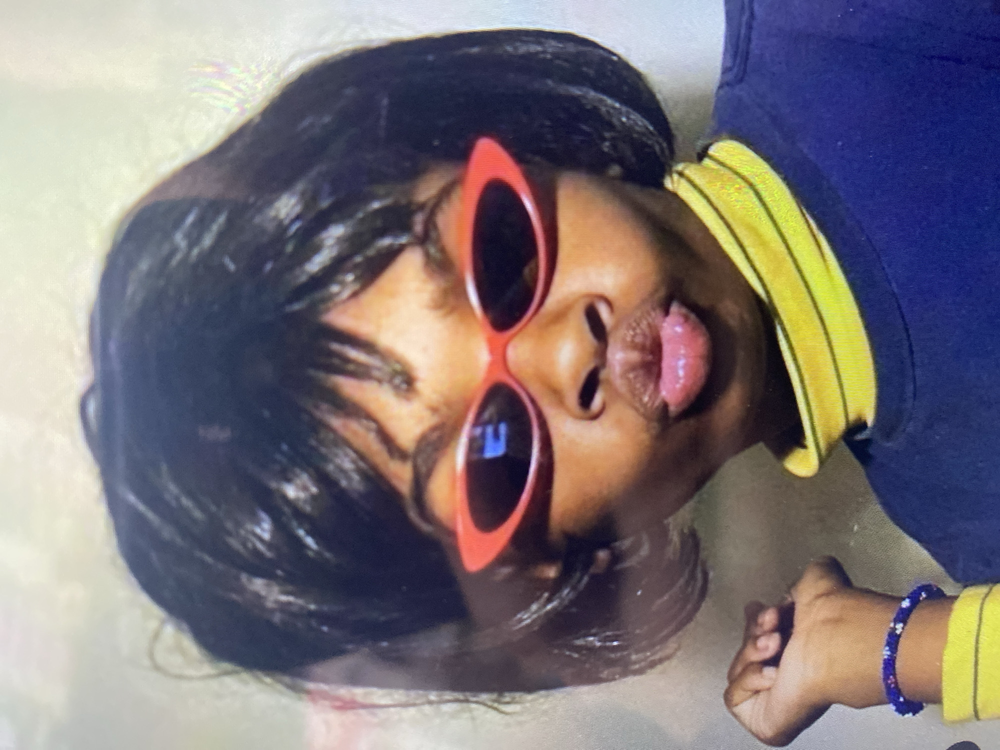 A close-up, waist-up shot shows a young person wearing a black wig styled in a bob, red cat-eye sunglasses, and a dark blue long-sleeved shirt with a yellow striped collar. The person's lips are pursed, and they are wearing a beaded bracelet on their left wrist. The background is a plain, light-colored wall. There is no visible indication of the location being in Ta', Indonesia.