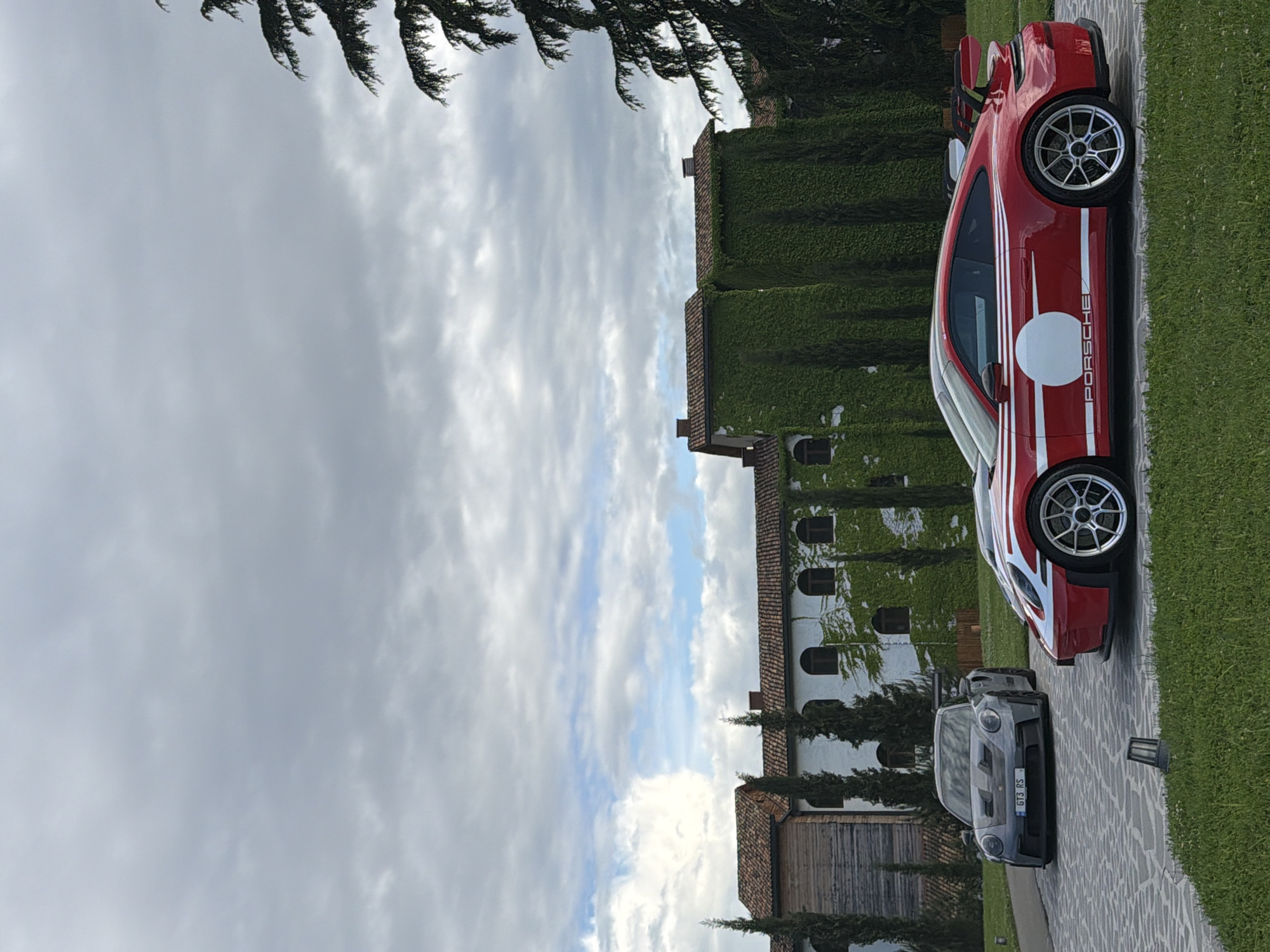 The image shows two Porsche GT3 RS cars parked on a stone driveway in Napareuli, Georgia. One Porsche is red with white stripes and a white circle, and the other is gray. A white building covered in ivy stands adjacent to the driveway. The sky is overcast with gray and white clouds. Evergreen trees are visible throughout the scene.