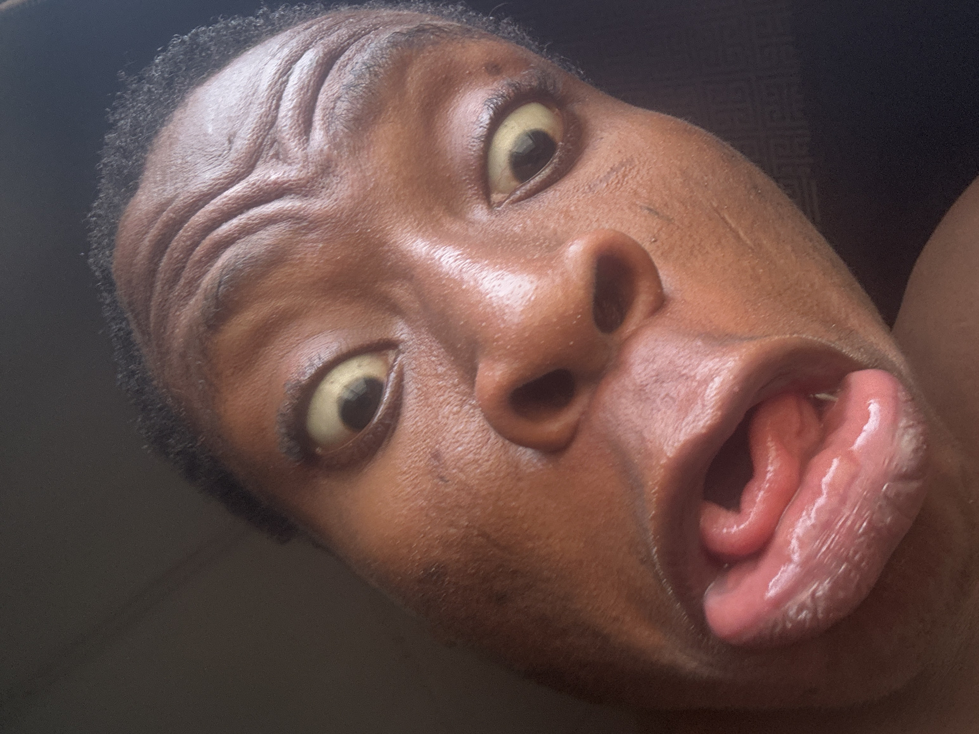 A close-up, high-angle shot captures the face of a person with dark skin, making an exaggerated and humorous expression. The person's head is tilted, showing the right side of their face more prominently. Their short, dark, curly hair is visible along the left side of their forehead.
The most striking features are their wide-open eyes, with a significant portion of the sclera visible, which appears distinctly yellowish. Their eyebrows are raised, causing prominent wrinkles across their forehead. The person's mouth is agape, revealing a large, pink tongue that is curled and contorted in an unusual way, extended outwards. The skin on their face appears somewhat shiny, suggesting moisture or oil. The overall expression conveys a sense of playfulness, surprise, or an intentional attempt to make a funny face.
The background is uniformly dark and out of focus, providing no specific location cues or details about the setting. There is no visible text in the image. The lighting appears soft and even, highlighting the contours of the face and the texture of the skin.