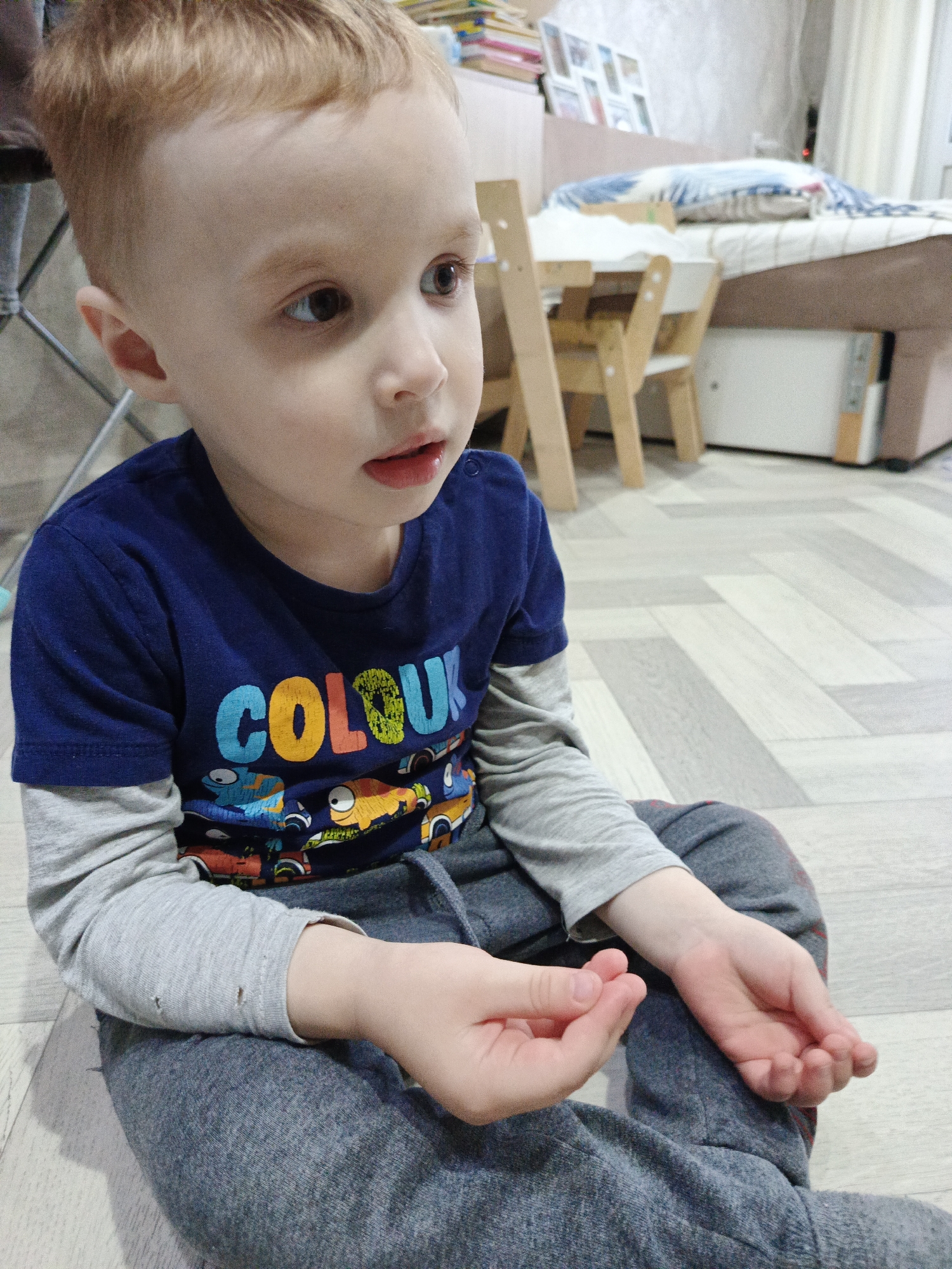 A young boy with reddish-blonde hair is sitting cross-legged on a light grey wood-patterned floor. He is wearing a blue t-shirt with the word "COLOUR" printed on it in large, colorful letters, and a grey long-sleeved shirt underneath. He is also wearing grey sweatpants. The boy is looking off to the right side of the image, with a slightly open mouth. His hands are in front of him, and he appears to be holding something small between his fingers. In the background, there is a small wooden table and chairs, a bed, and a cabinet. The location is a room within an unknown building, in the city of Unknown, Unknown.