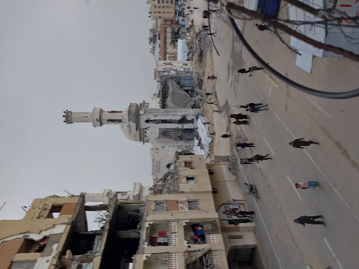 The image displays a scene of extensive urban destruction in Khan Yunis, Palestinian Territory, under an overcast sky. A large mosque, identifiable by its dome and a prominent minaret, stands in the midground, exhibiting severe structural damage, including significant impact marks, a scarred dome, and a collapsed section at its base. To the left, a multi-story building, appearing to be residential, is heavily damaged; large portions of its facade are torn away, exposing internal rooms, rebar, and debris. Clothes are visible hanging on partially intact balconies on this structure.
On the paved street below, approximately 20-30 individuals, predominantly adults, are observed walking or standing. The street features visible white lane markings. An overturned motorcycle is present on the sidewalk adjacent to the damaged residential building. To the right of the street, the area is covered in large piles of concrete rubble and the remnants of completely destroyed structures. Further in the background, additional damaged buildings are visible. Overhead utility lines are partially visible in the bottom right foreground.