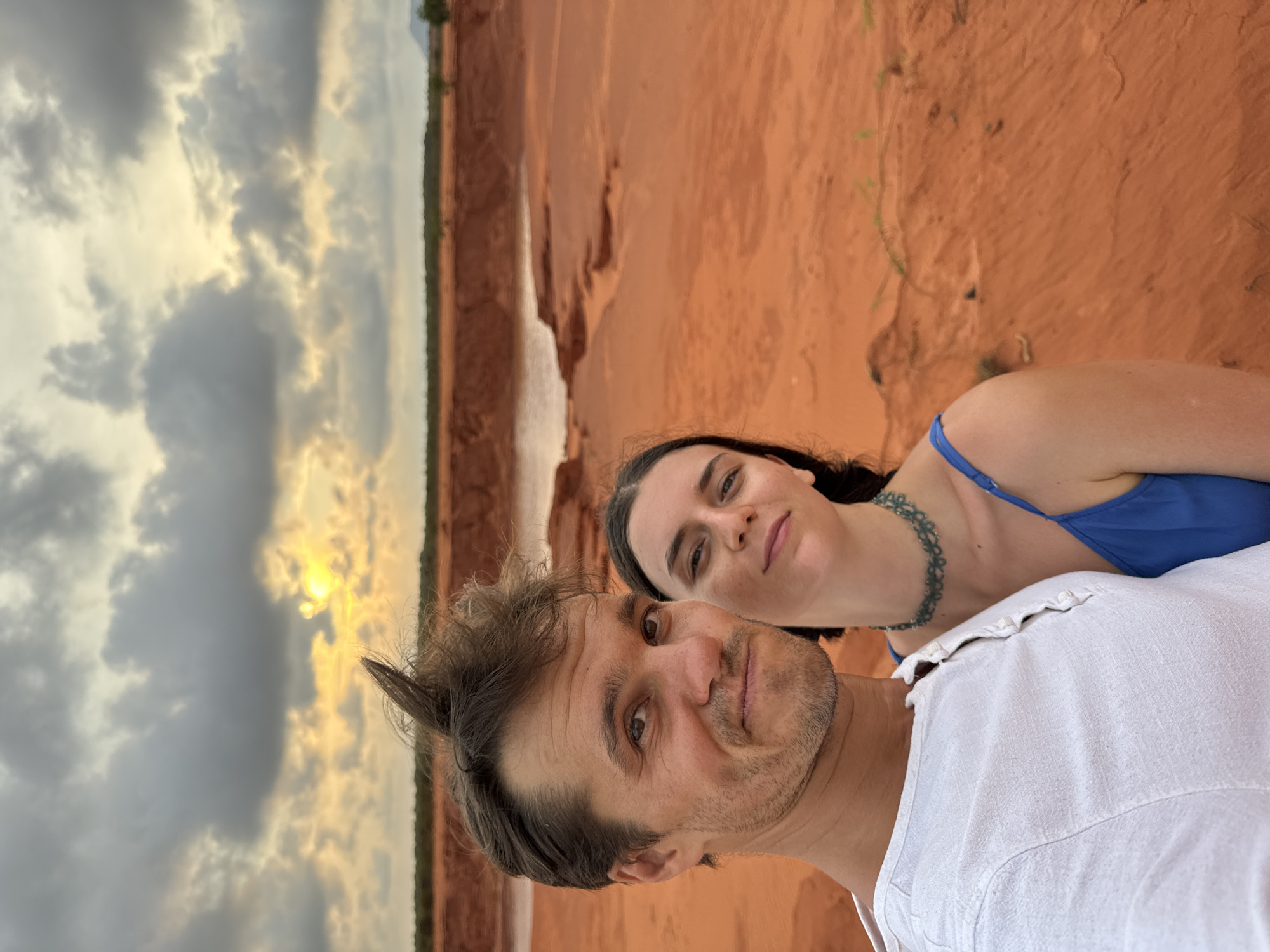 A selfie shows a Caucasian male and female in Mui Ne, Vietnam. The male is in the foreground, wearing a white shirt. The female, positioned behind him, is wearing a blue top and a green choker necklace. In the background is a red canyon landscape, and a cloudy sunset sky.