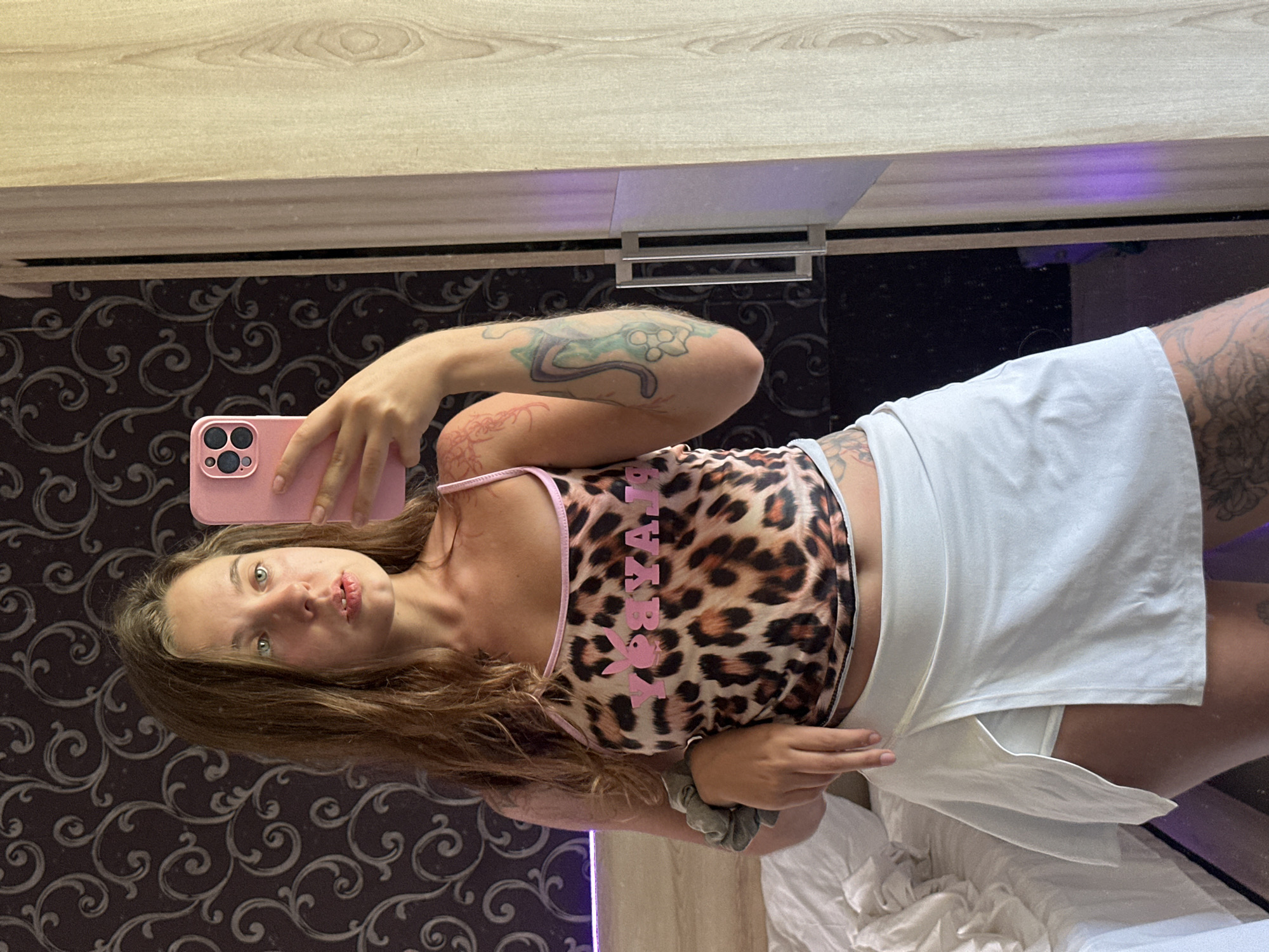 A young woman with long, wavy brown hair is standing in a bedroom and taking a selfie in a mirror. She is wearing a leopard print crop top with pink trim and the word "PLAYBOY" printed on it in pink letters, and a light-colored, short skirt with a slit. She has tattoos on her arms and legs. The background shows a bed with white linens, a dark patterned wallpaper, and a wooden closet. A pink phone is held in her right hand, and she is looking at her reflection. The lighting is bright and natural.