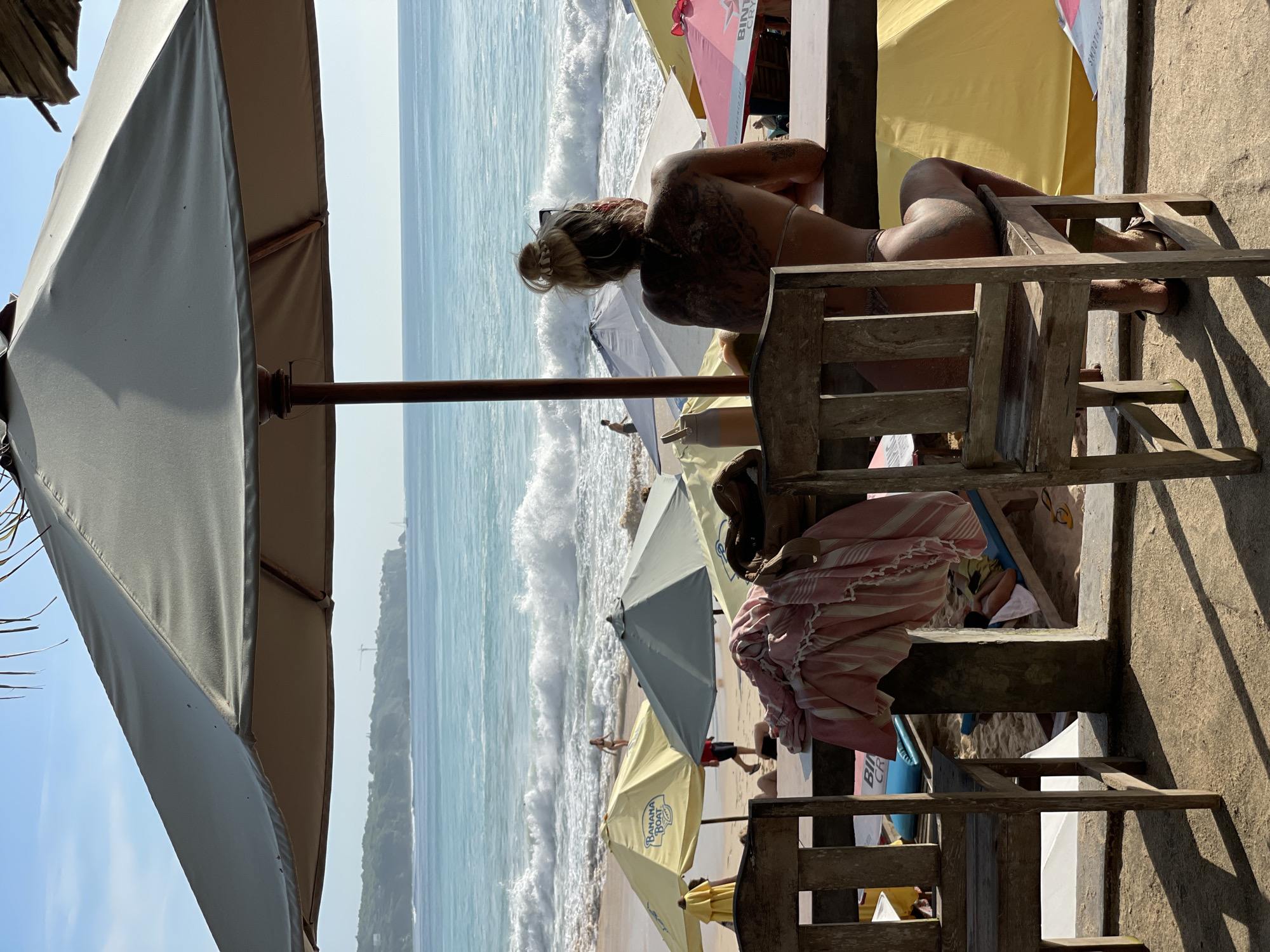 This image captures a vibrant beach scene in Kuta Selatan, Indonesia, from an elevated perspective overlooking the shore and ocean. The time of day appears to be sunny, likely late morning or early afternoon, casting distinct shadows.
In the midground, a person with blonde hair, styled in a bun, is seen from behind, leaning forward on a wooden railing or counter. Their back is covered in sand and possibly a large tattoo. They are wearing a bikini top and looking out at the crashing waves of the blue-green ocean. A wooden chair is visible next to them.
The immediate foreground on the left is dominated by a large, beige beach umbrella, partially obscuring the view of the sky. On the right, wooden structures, including a railing and parts of chairs, are visible, suggesting a seating area or balcony.
Further down on the sandy beach, numerous other people are scattered, some walking near the waterline, others relaxing under various beach umbrellas. These umbrellas come in different colors, including yellow, light blue/grey, red, and off-white. One red umbrella prominently displays the text "BINTANG" and "SMOOTH." Another yellow umbrella features "BANANA BOAT" text.
The ocean beyond the beach shows white-capped waves breaking on the shore. In the far distance, a lush green headland or hill is visible under a clear, bright blue sky. A blue surfboard is also partially visible on the beach. The overall scene depicts a relaxed, sunny day at a popular beach destination.