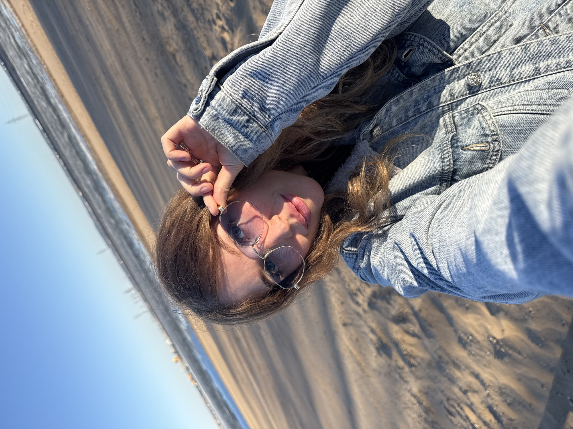 A person wearing sunglasses and a blue denim jacket is lying on a sandy beach, possibly in Noviy, Russia. The background shows a clear blue sky and the sea. The person's head is oriented towards the top-left corner of the image. The sand appears to be slightly wet, indicating its proximity to the sea.