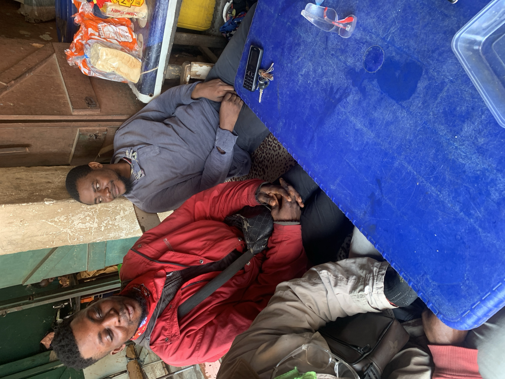 Two men are seated at a table in what appears to be an outdoor market stall in Shagamu, Nigeria. The man on the left is wearing a red jacket and looking down and to his left, with his mouth open as if speaking. He has a red and white patterned bandana around his neck and a black crossbody bag. The man on the right is wearing a grey long-sleeved shirt and looking to his left with a slight smile. On the blue plastic table in front of them are a mobile phone, a set of keys, a clear plastic container, and a pair of red-rimmed glasses. Behind them, a shelf holds several loaves of bread in plastic packaging. The background shows a rustic building facade with wooden doors and a green metal structure.