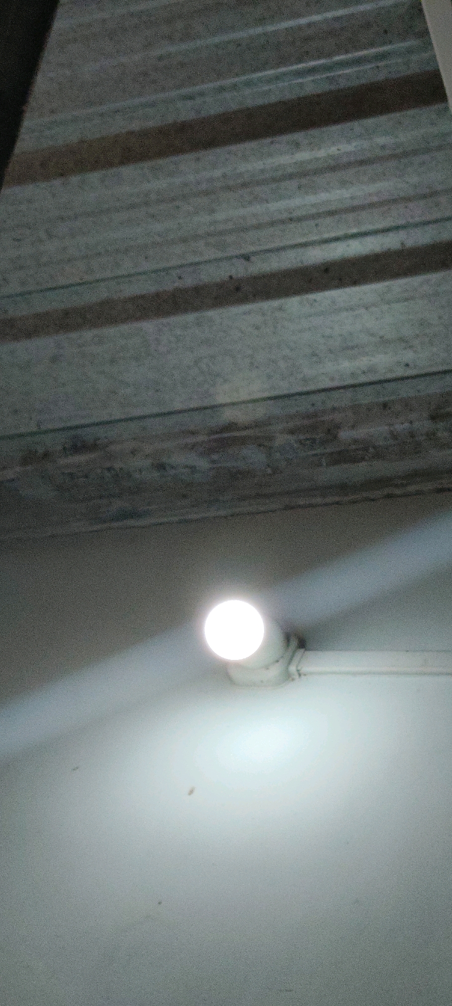 The image shows a white LED bulb emitting light, mounted on a white conduit pipe. The pipe is affixed to a textured light-colored wall, which is illuminated by the bulb. Above the wall, a corrugated metal ceiling is visible, appearing weathered and dusty. The lighting suggests an indoor setting, possibly a basement or an unfinished area. The scene is devoid of any people or specific landmarks that would indicate a precise location within Belagavi, India. The composition is a close-up of the light fixture and the surrounding surfaces.