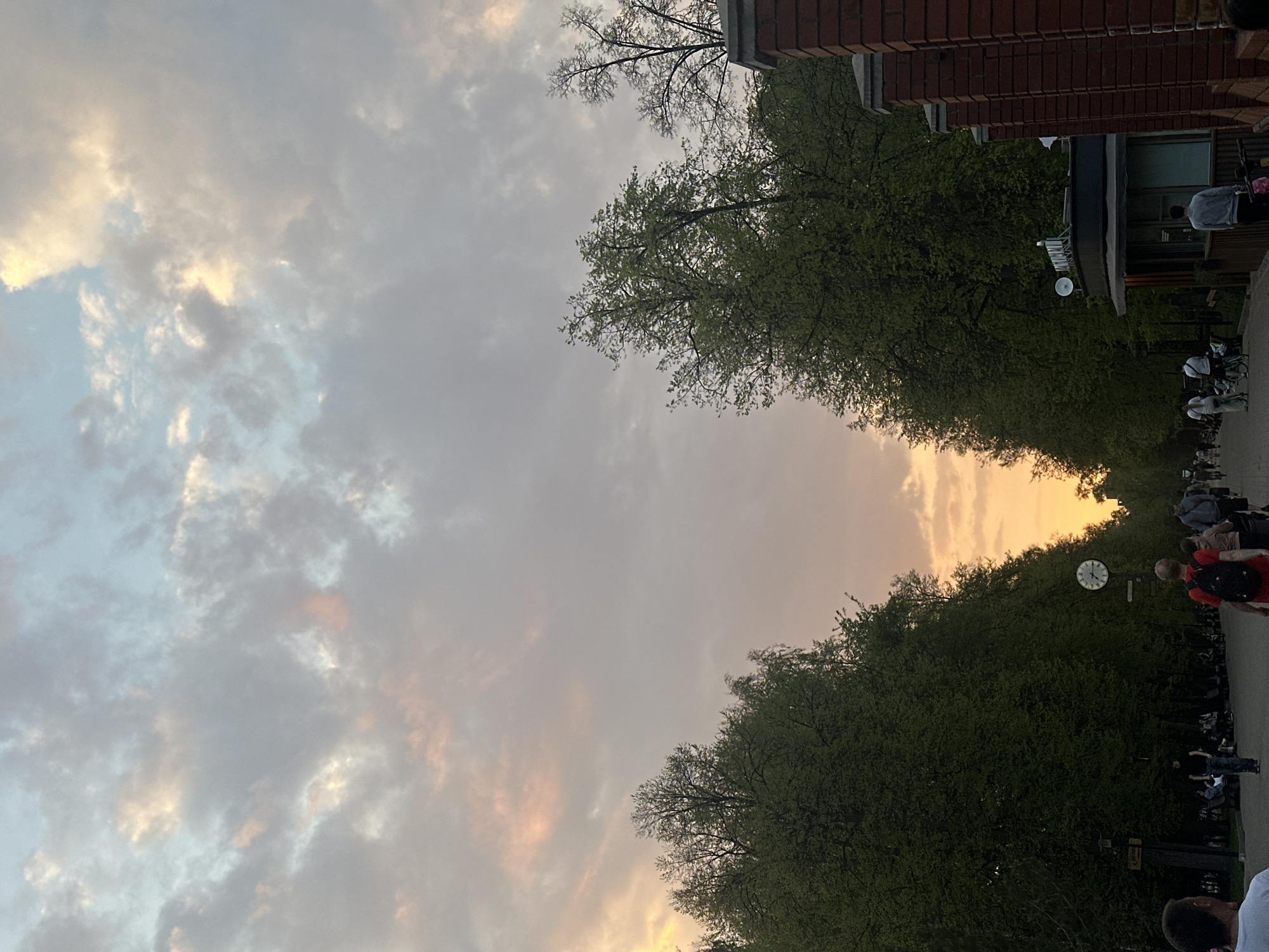 The image shows a street scene in an urban environment. The sky is filled with clouds, tinted with the colors of the setting sun. Rows of trees line the street, meeting overhead to create a tunnel-like effect. On the right side of the frame, there is a brick building and a building with a flat roof. Pedestrians and parked strollers can be seen along the sidewalk. A clock is mounted on a pole, indicating the time.