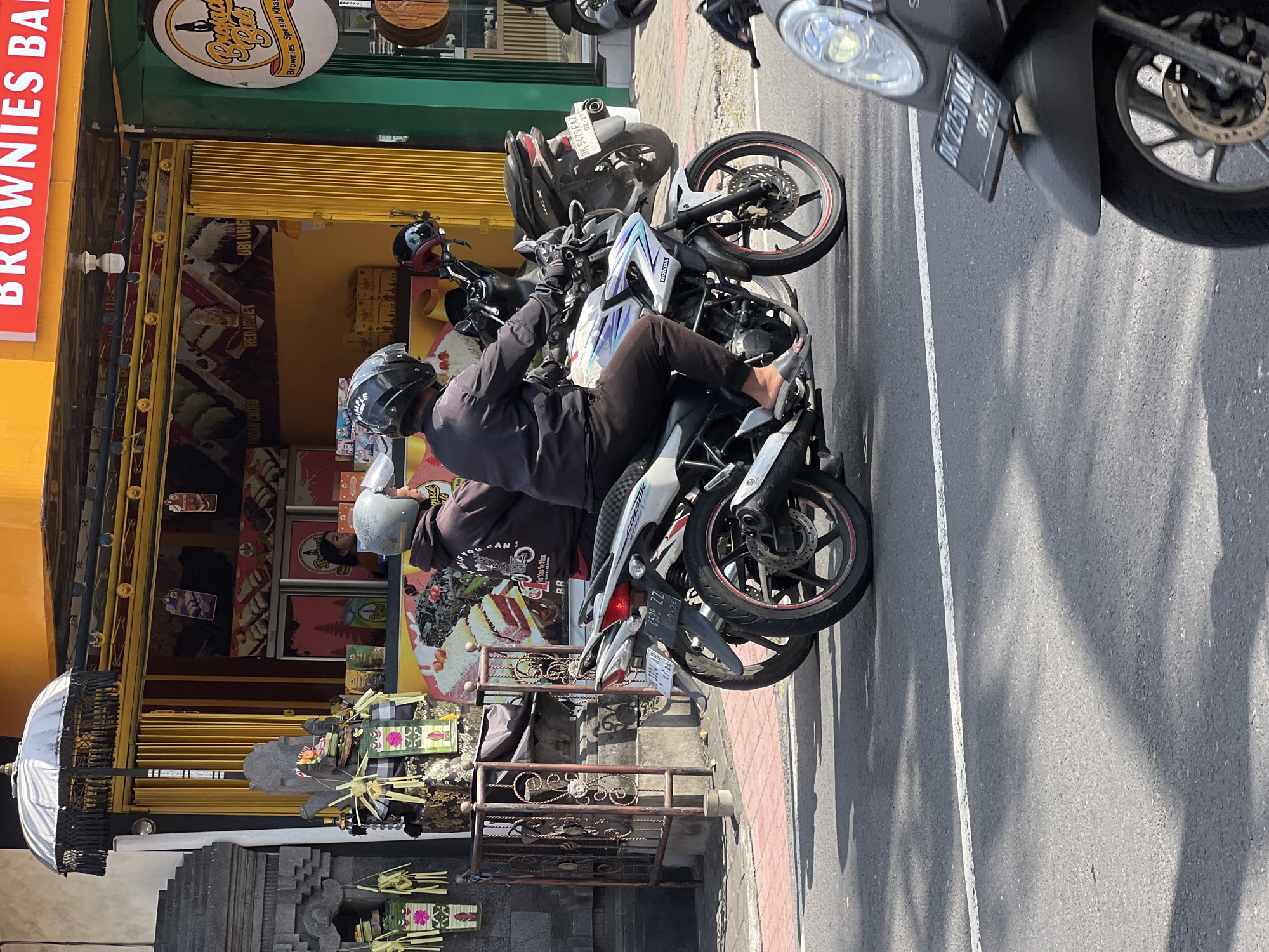 Two individuals are riding on a white and blue motorcycle on a street in Denpasar, Indonesia. The rider in front is wearing a black helmet and a black hooded sweatshirt. The passenger behind is wearing a grey helmet and a grey hooded sweatshirt. They are traveling on an asphalt road with a white line marking. In the background, there is a building with a yellow awning and red lettering that says "BROWNIES BA." Adjacent to this is a green storefront with a circular logo. There are other motorcycles parked or in motion on the street, and some decorative elements, possibly religious or cultural, are visible on the left side of the image.