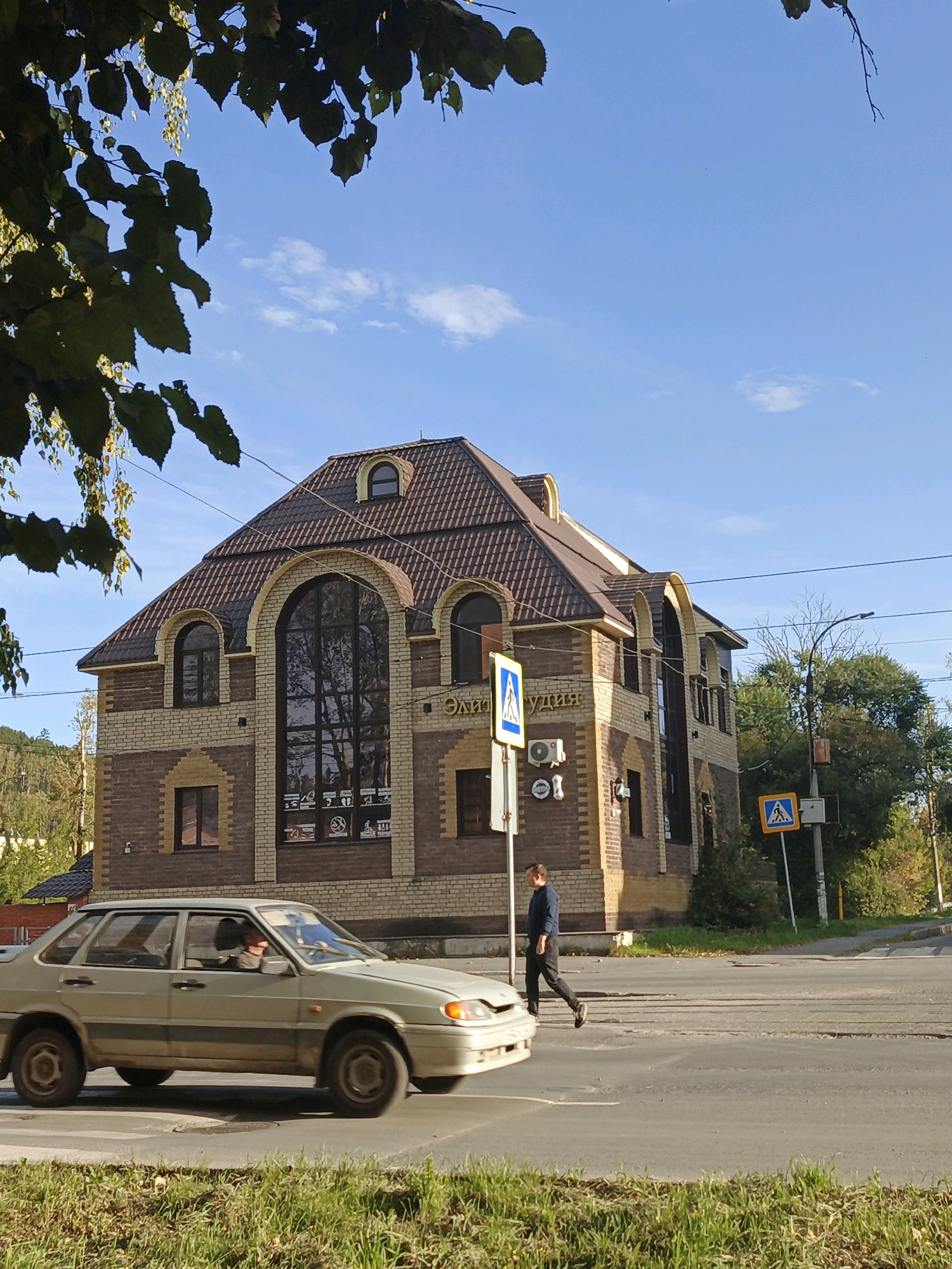 In Zlatoust, Russia, a light-colored sedan drives across a crosswalk. A person in a dark blue shirt and black pants is walking across the same crosswalk. Behind them, a brick building with a brown roof features large arched windows. The sky is blue with scattered white clouds. The scene is framed by dark green leaves from a tree in the foreground.