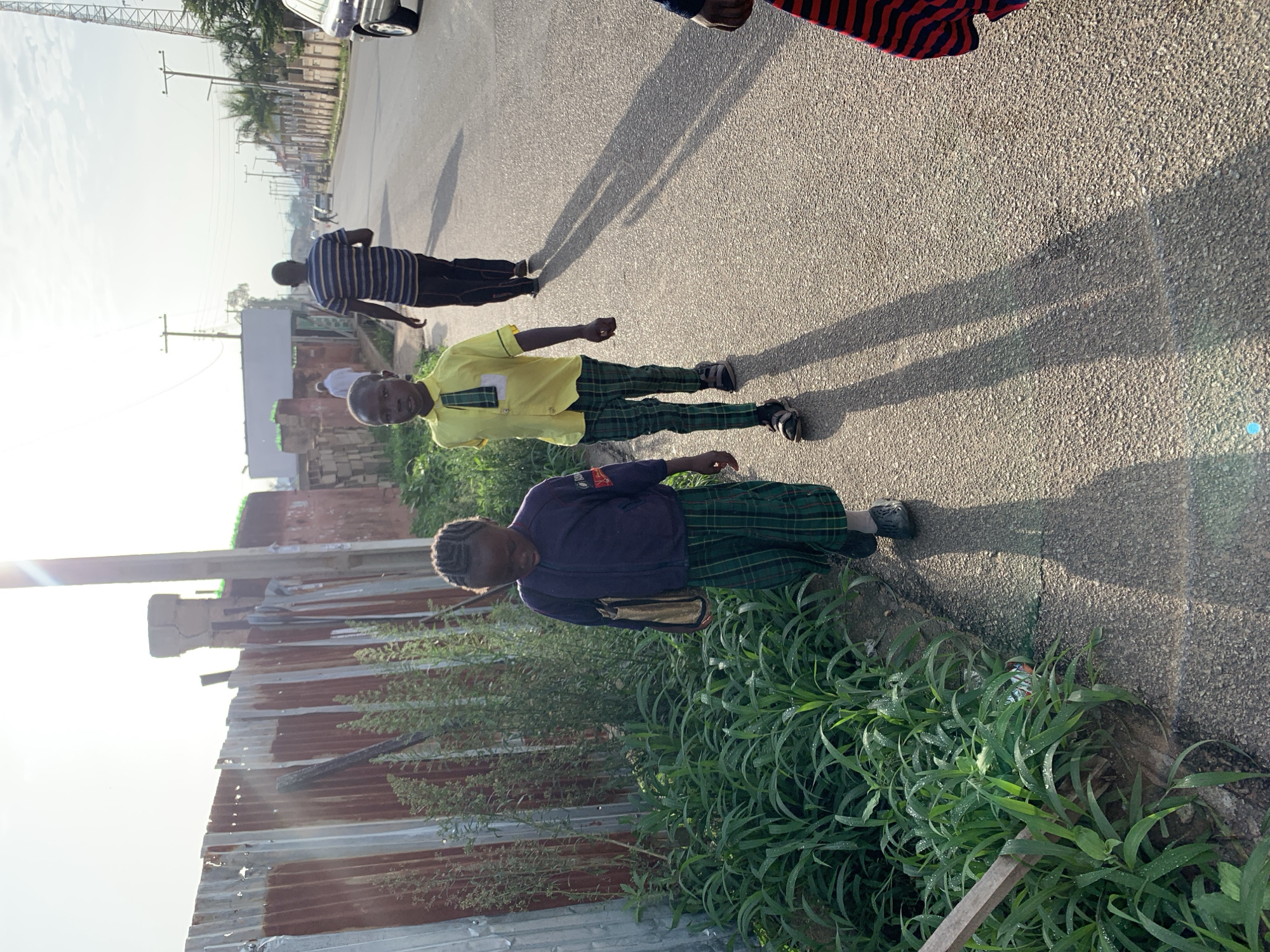 In Jos, Nigeria, two young children, a boy and a girl, walk on a paved road next to a corrugated metal fence. The boy, in the foreground, wears a yellow shirt and green plaid pants. The girl, to his left, wears a dark jacket and matching green plaid pants, carrying a tan bag. Both children cast long shadows on the road, indicating late afternoon sun. A man in a striped shirt walks further down the road, along with other pedestrians and vehicles in the distance, suggesting a public street in a developed area. The environment appears to be an urban setting with some overgrowth of green vegetation along the roadside.