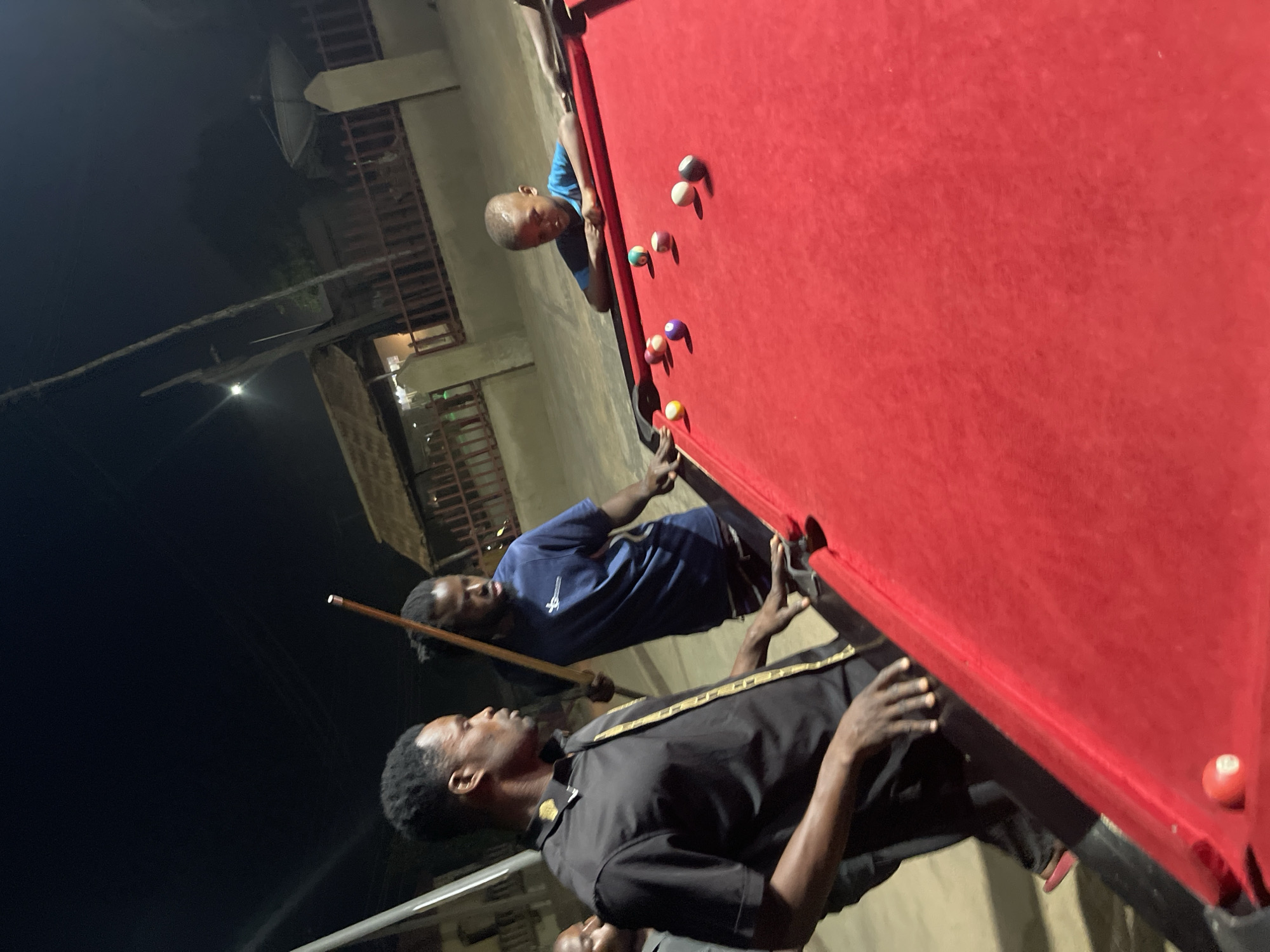 This vertical image depicts an outdoor night scene in Unguwar Gabas, Nigeria, centered around a game of pool. The main subjects are at least three men gathered around a vibrant red-felted pool table.
One man, wearing a blue short-sleeved shirt, is positioned at the far end of the table, leaning over the red felt, appearing to concentrate on the arrangement of the pool balls. Another man, dressed in a black short-sleeved shirt with gold trim along the placket, stands closer to the foreground, holding a pool cue stick in his left hand, ready to play or observing the game intently. A third man, also in a dark shirt, is partially visible near the table's edge in the lower part of the frame.
Numerous pool balls of various colors – including white, black, purple, yellow, and striped balls – are scattered across the bright red surface of the table. One red and white ball is visible near the table's corner.
The time of day is clearly night, as indicated by the dark, unlit sky overhead. Artificial lights, though dim, illuminate the immediate area around the pool table and cast shadows. In the background, indistinct outlines of buildings with railings and what appears to be a large satellite dish are visible, suggesting a residential or commercial setting. The men appear focused on the game. No explicit text is visible on any objects or clothing.