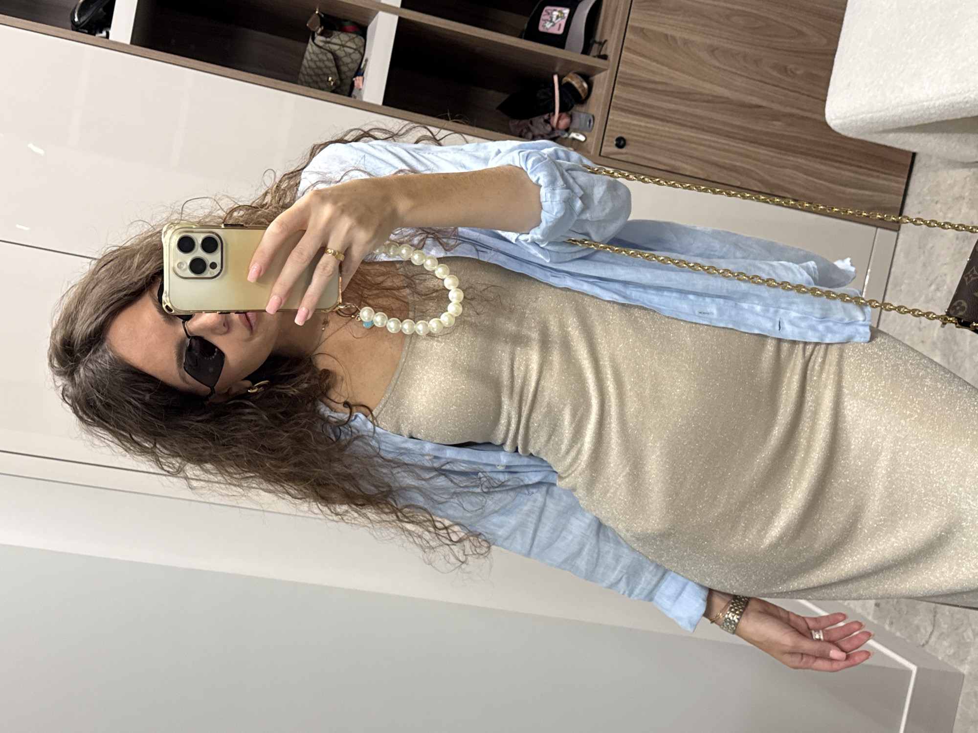 A woman is taking a mirror selfie in what appears to be a closet or dressing area. She has long, curly brown hair and is wearing dark, angular sunglasses, a light blue linen shirt unbuttoned and worn over a sleeveless, shimmering champagne-colored midi dress. She is also wearing a pearl necklace and a gold ring on her right hand. Her left arm is bent, holding a gold iPhone with a clear case. A gold chain purse strap is visible over her left shoulder, with the purse hanging down. In the background, there are wooden shelves with various items including bags, hats, and other accessories. The location is identified as Khimki, Russia.