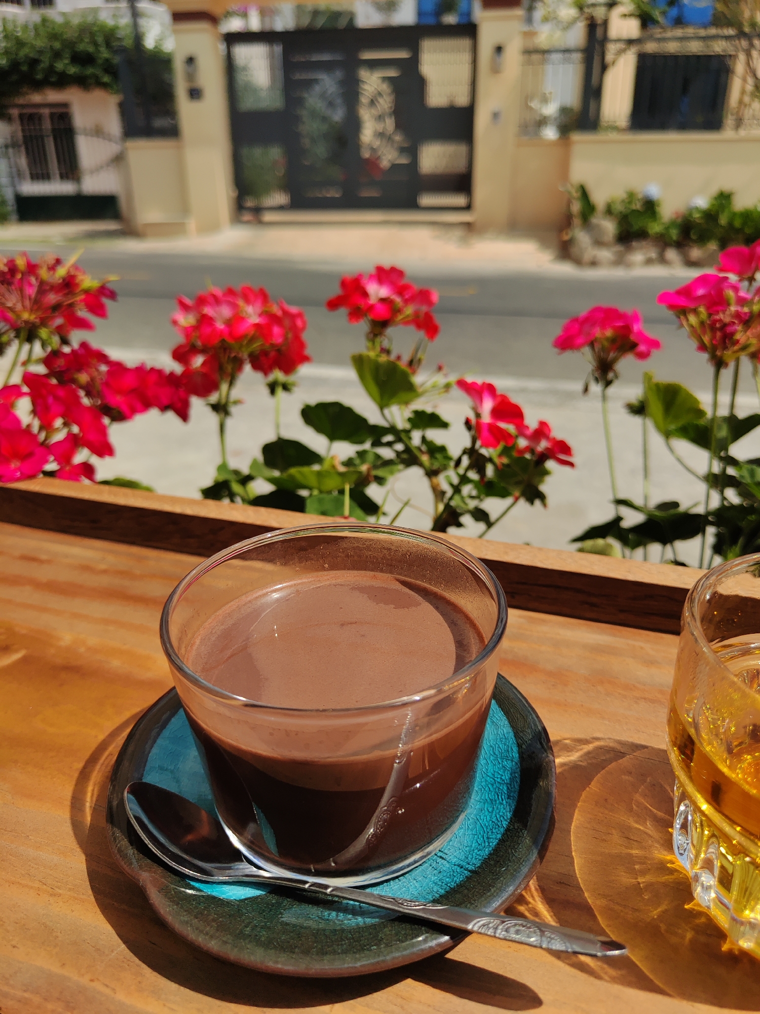 The scene depicts a close-up of a wooden table with two glasses, set against a blurred background. The foreground is dominated by a clear glass filled with a dark brown liquid, likely chocolate milk, resting on a turquoise saucer with a silver spoon placed on top. To the right, another glass is partially visible, filled with a yellow liquid. The background is blurred but suggests an outdoor setting with red geranium flowers in a planter. Beyond the flowers is a street with buildings, including a house with a dark gate. Based on the presence of the geranium flowers, commonly grown in the area, and the style of architecture visible, it can be concluded that the location is in the city of Lâm Viên - Đà Lạt, Vietnam. There are no people visible in the scene. The focus is on the drinks and the serene outdoor environment.