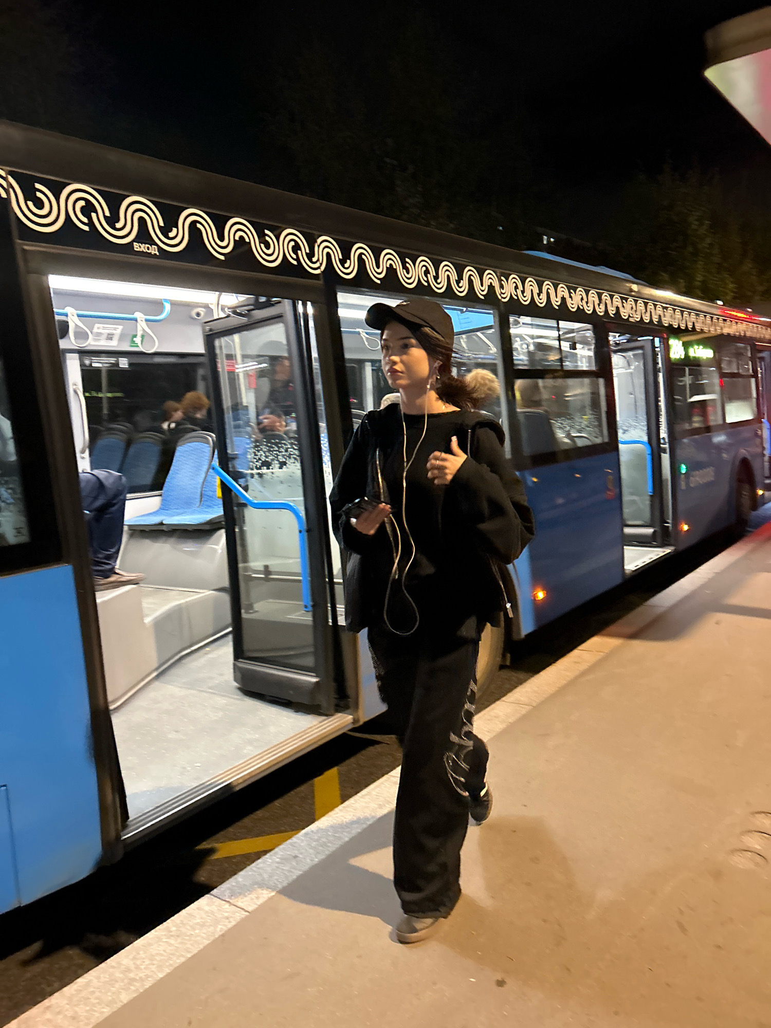 A young woman wearing a black baseball cap and black athleisure clothing walks towards the viewer on a paved sidewalk next to a blue public bus with its doors open. The bus has a decorative wavy illuminated trim along its roofline and the word "ВХОД" (Entrance) above the front door. Inside the bus, blue seats are visible. The scene appears to be at a bus stop at night, illuminated by streetlights and the bus's interior lights, suggesting a location in Moscow, Russia, given the Cyrillic lettering on the bus.