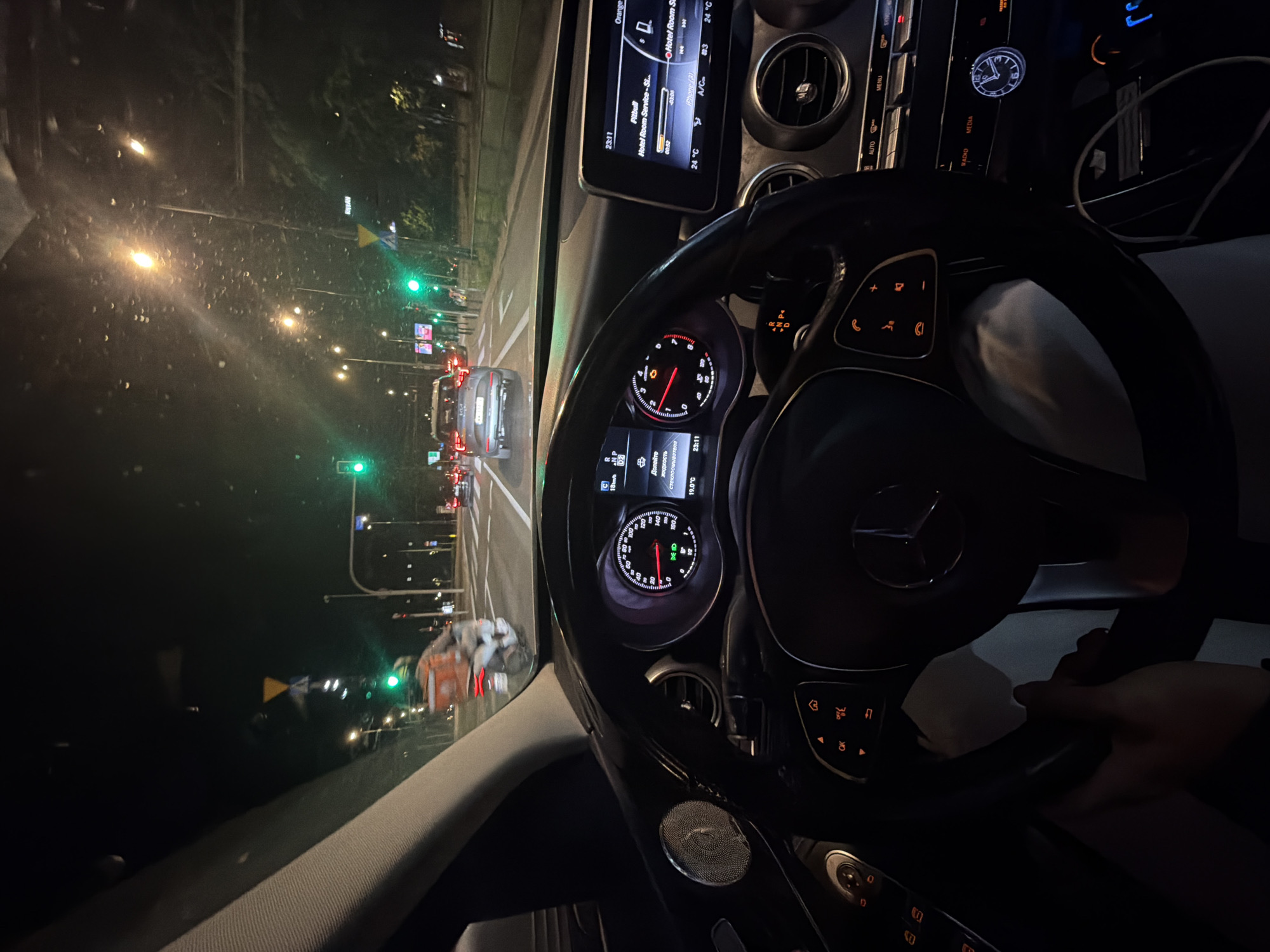 The image depicts the interior of a car at night in what appears to be an urban setting in Unknown, Unknown. The perspective is from the driver's seat, looking out at a street with multiple traffic lights, all currently green. There are several cars ahead, with their tail lights illuminated. The car's dashboard is visible, with the steering wheel prominently displayed in the center. Information displayed on the car's screen shows a song playing ("Hotel Room Service"), an active navigation, and the time (23:11). The temperature is 24°C, and the air conditioning is on. The speedometer is visible and shows the car is currently stopped. The gear is in "Drive." A message on the instrument panel indicates a need to refill the windshield washer fluid. A hand is partially visible on the right side of the steering wheel.