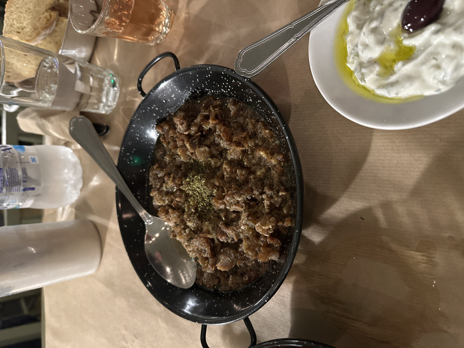 An overhead shot captures a casual dining experience at a table, likely in a Greek restaurant in Nea Erithrea, Greece. The table is covered with brown paper, and the scene is illuminated by warm, artificial light, casting soft shadows.
The focal point is a black pan, possibly cast iron, filled with a dark, savory chopped meat or offal dish, generously garnished with dried herbs, likely oregano. A silver spoon rests in the dish. To the right, a white, boat-shaped bowl holds creamy tzatziki, drizzled with olive oil and topped with a single black olive. Another silver spoon rests on the edge of this bowl.
Various other items are visible on the table: several slices of rustic bread in a basket, a clear glass of water, and a glass containing a rose-colored beverage, possibly rosé wine. A plastic water bottle, partially visible on the left, has "APOA" and "NATURAL MINERAL WATER 2L" printed on its label in Greek and English, confirming the setting's context. A rolled napkin or paper towel is also partially visible. The arrangement suggests a meal is either about to be enjoyed or is currently underway.