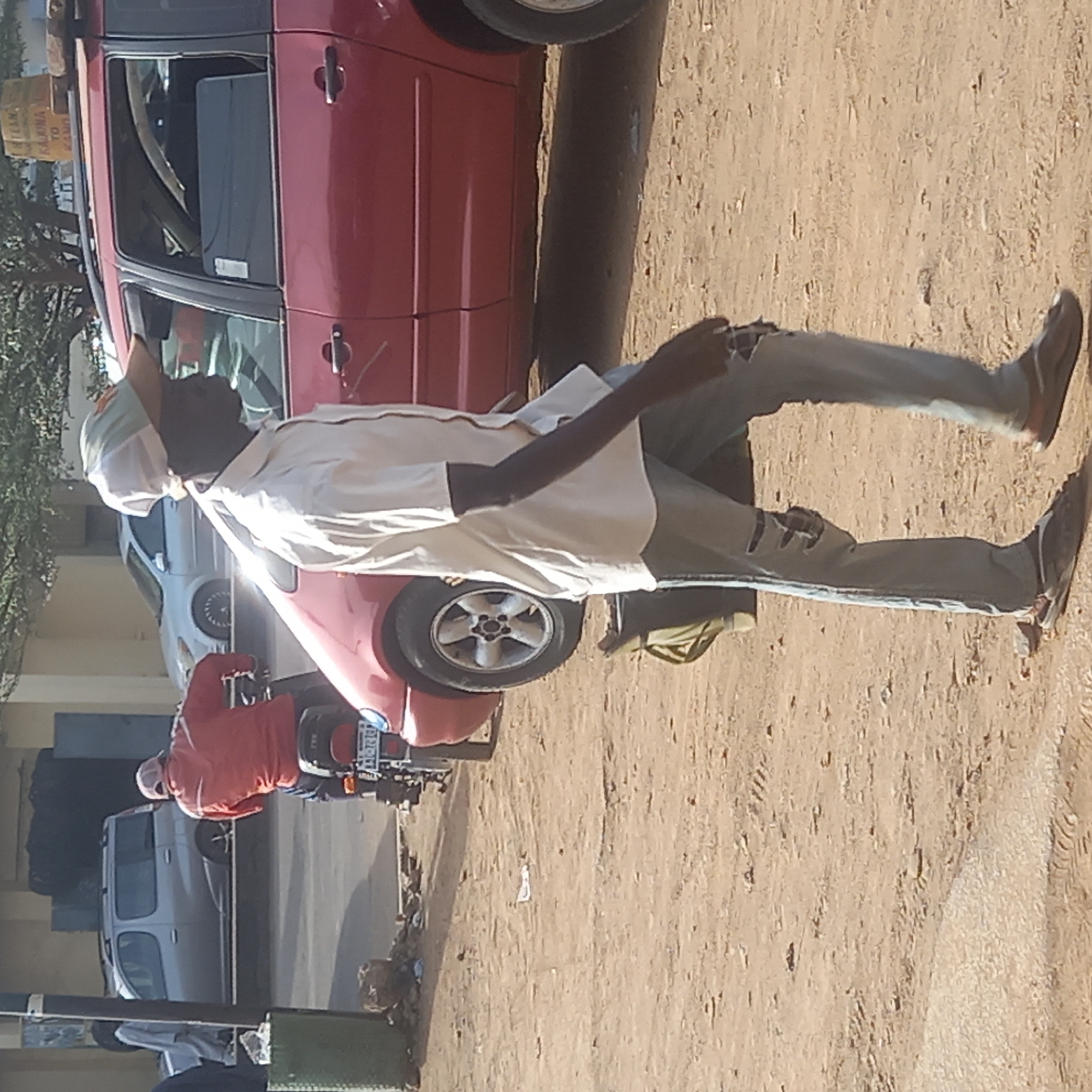 The image captures a daytime scene on an unpaved, dusty ground next to a road, likely in Sardauna, Nigeria. The main subject is a dark-skinned man, seen from the waist down, wearing a light-colored long-sleeved shirt, distressed light blue jeans with rips at the knees, and dark shoes. He also has a white or light-colored head covering or mask. He appears to be walking on the unpaved surface.
To his right (image left, if upright) is a red car, possibly a sedan or hatchback, parked or stopped. On its roof rack, a cardboard box is visible with handwritten text, which appears to read "TALEN KAFINA TO KANO", suggesting a destination for a journey. Behind the red car and the main subject, another person in a red jacket is seated on a motorcycle or scooter, facing away from the camera. Further in the background, a silver or grey car is parked on what seems to be a paved road, with buildings and trees visible behind it. The lighting indicates bright sunshine. The overall atmosphere suggests a busy, utilitarian street or market area. A dark green object, possibly a pole or bin, is partially visible in the lower left corner.