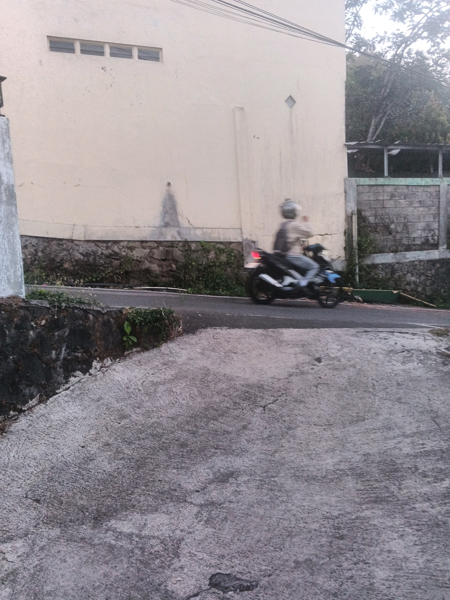 The image depicts a street scene in Keji, Indonesia, characterized by a light-colored building with horizontal rectangular windows on the left, and a gray stone wall topped with a greenish border and brickwork on the right. A motorcycle with a rider wearing a helmet is captured in motion on the asphalt road that runs horizontally across the mid-ground. The road is bordered by a dark, rough stone embankment on the left and vegetation. In the foreground, a textured gray concrete surface slopes upwards from the bottom of the frame. The lighting suggests it is daytime, possibly late afternoon.