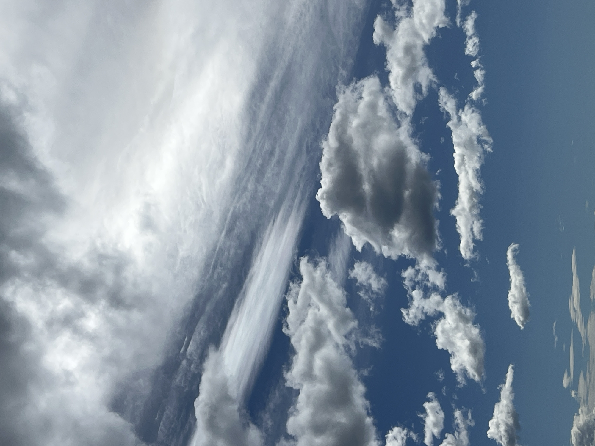 The image presents a daytime view of a sky filled with diverse cloud formations. On the left side, there are dense, grey clouds suggesting possible overcast conditions. Towards the center, there are streaks of cirrus-like clouds layered with cumulus clouds, with a prominent vertical streak. The right side shows patches of blue sky punctuated by small, scattered cumulus clouds. No people or distinct objects are identifiable in the image. The environment is solely atmospheric. There is no indication of Prijevor, Montenegro, or any terrestrial elements.