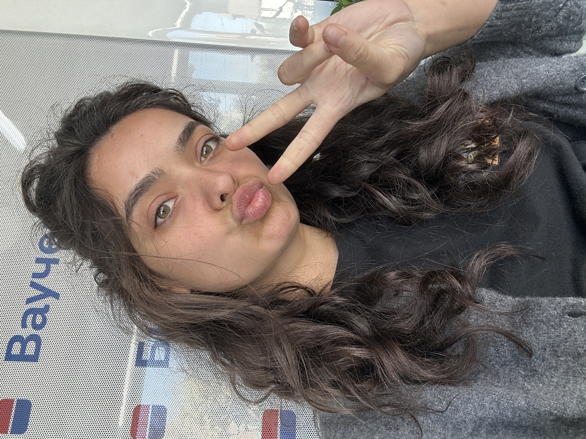 A young woman with long, dark, wavy hair is taking a selfie. She is wearing a grey cardigan over a black t-shirt. Her lips are pursed in a kiss, and she is holding up two fingers in a peace sign. Behind her is a perforated wall with blue and white lettering that reads "Вауче". The image appears to have been taken indoors.