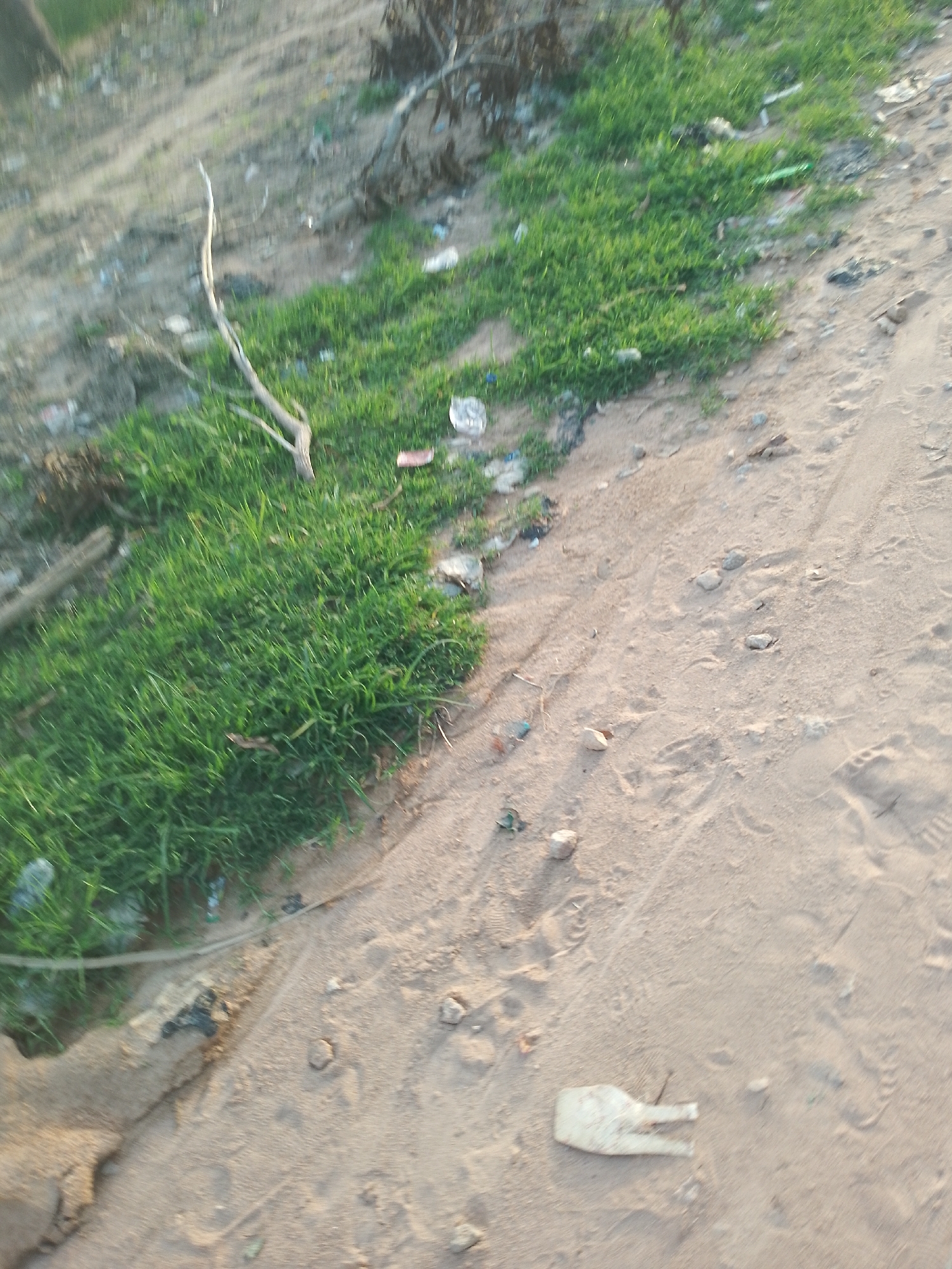 The image shows a dry, sandy area with patches of green grass. Several pieces of debris, including plastic bottles and other refuse, are scattered across the scene. A piece of light-colored plastic, possibly a discarded shoe or part of one, lies on the sand. Footprints are visible in the sand, indicating human activity. The overall impression is one of an unkempt outdoor environment, possibly a roadside or undeveloped area in Jalingo, Nigeria. The lighting suggests it might be late afternoon or early morning.