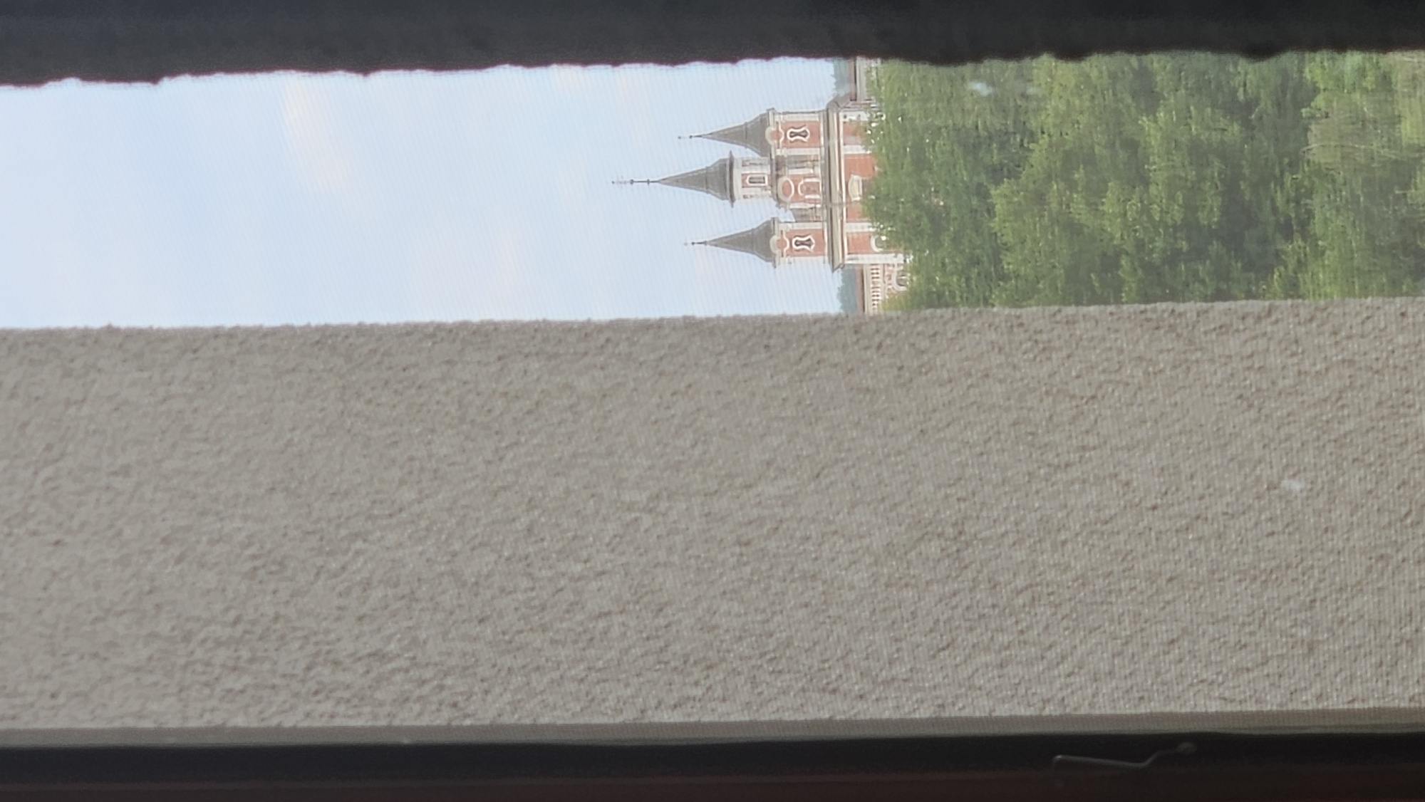 The image depicts a view through a window frame, with a rough textured wall on the left and a dark curtain on the right. In the distance, a building with distinctive red and white architecture and multiple spires is visible, surrounded by lush green trees under a pale blue sky. This building is the Slovak Academy of Sciences building in Prešov, Slovakia.