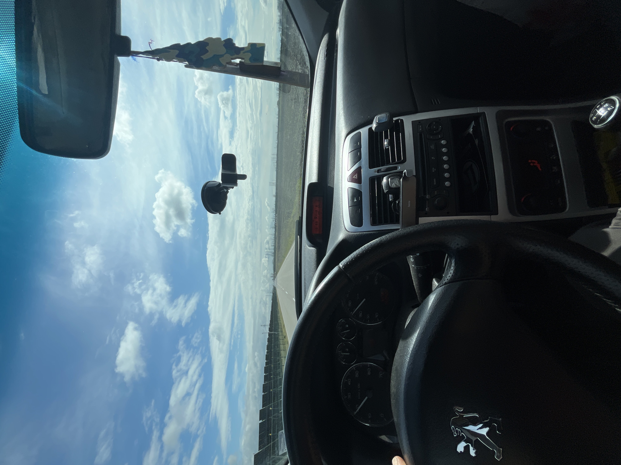This image presents a first-person view from the driver's seat of a Peugeot vehicle, observing a straight road in the vicinity of Dalgo Pole, Bulgaria. The scene is illuminated by daylight under a blue sky with scattered white cumulus clouds.
In the foreground, the black steering wheel, featuring the silver Peugeot logo, occupies the lower-left quadrant, with a hand partially visible gripping its left side. The instrument cluster behind the steering wheel shows a speedometer indicating approximately 60 km/h. Above the central console, an orange digital display is visible. The central console itself contains a car radio, climate control buttons, and air vents, with the red hazard light button illuminated. A portion of a manual gear shift lever is visible in the lower-right. A black suction-cup mount is affixed to the upper-left of the windshield, and a camouflage-patterned air freshener hangs from the rearview mirror, positioned in the upper-right of the windshield.
Outside the vehicle, a straight asphalt road extends centrally into the distance, marked by a dashed white center line and a solid white line on the right shoulder. To the left of the road, a long, linear installation of dark solar panels is present. To the right, a large, flat, tilled field stretches towards the horizon. In the distant background, industrial structures, possibly a power plant with cooling towers or chimneys, along with several power transmission towers, are visible against the sky.