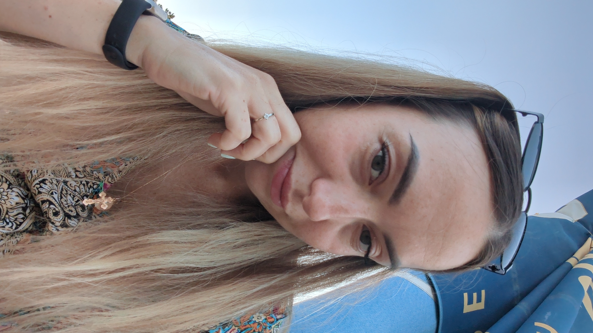 A woman with long blonde hair and sunglasses perched on her head is shown in a close-up selfie outdoors. She is wearing a colorful patterned top and a cross pendant. Her left hand is resting on her chin, and she is wearing a ring on her left ring finger. A black fitness tracker is visible on her left wrist. A portion of a blue umbrella with the letter "E" visible on it is in the background. The sky is clear and blue. The overall scene suggests a sunny day, possibly at a beach or park.