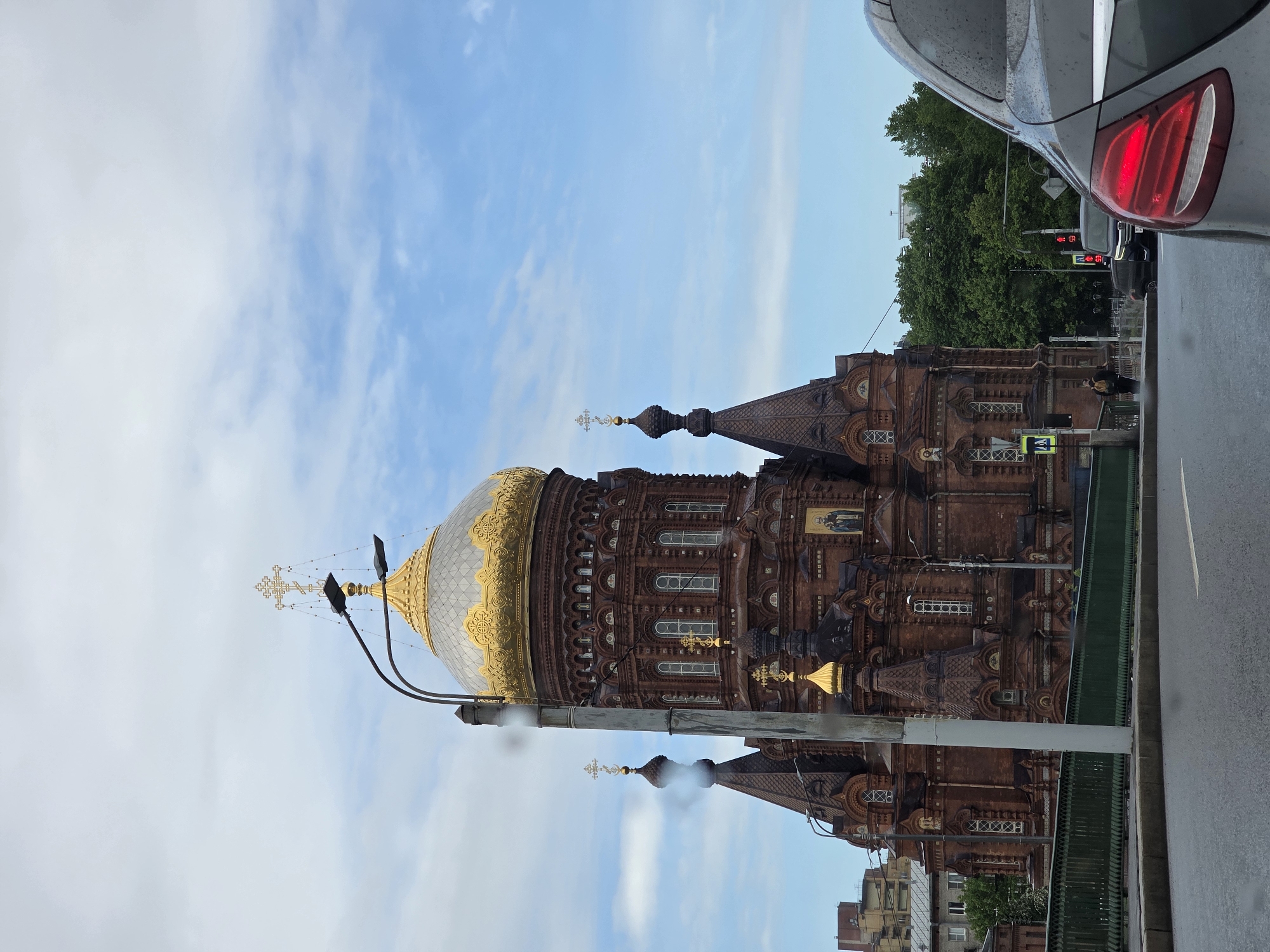 Here's a detailed description of the image:
**Scene:** An urban scene featuring the Church of the Savior on Blood in Saint Petersburg, Russia. The church's distinctive architecture is prominent, with its multi-colored onion domes and intricate brickwork. A golden dome is in the left of the image.
**Environment:** The sky is blue with some scattered clouds. The street is wet, suggesting recent rain.
**Objects:** A car is partially visible on the right side of the image, the tail light of the vehicle is visible. A traffic light is visible behind the car. There is a street lamp is in the image. The image is taken in portrait mode, with the top of the image being on the left side of the image.