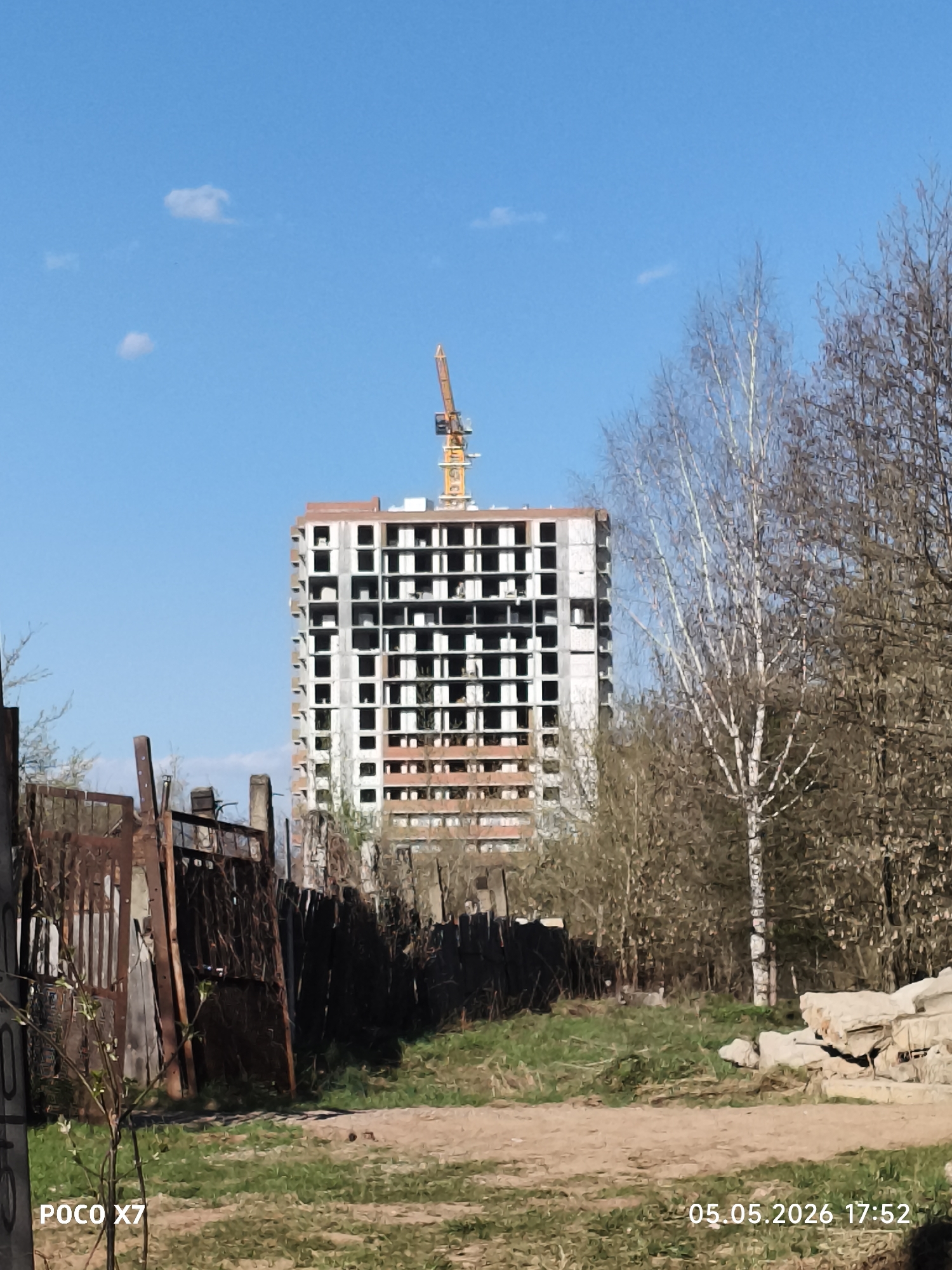 The image, dated May 5, 2026, at 17:52, depicts an unfinished high-rise building under construction in what appears to be a partially developed or neglected area. The building, situated against a clear blue sky with scattered clouds, is a reinforced concrete structure with many unfinished window openings. A yellow crane is attached to the top of the building. In the foreground, there is an unkempt area with a dirt path, overgrown grass, and a dilapidated, rusty fence. To the right of the frame, bare trees and concrete debris suggest a neglected or undeveloped surrounding. The camera is a POCO X7. Based on the location, this image is most likely taken in the city of Chaikovsky, Perm Krai, Russia.