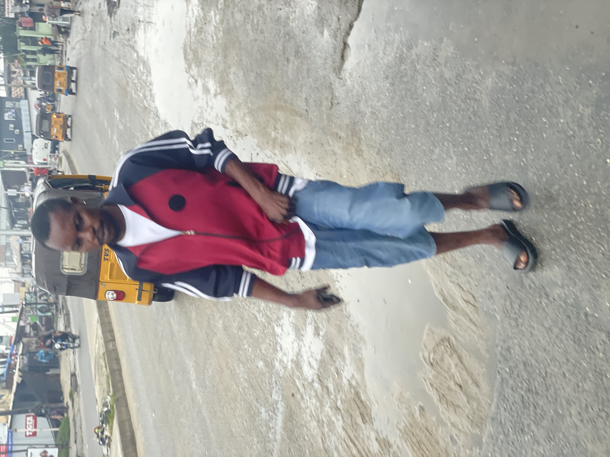 A medium shot shows a dark-skinned man standing on a wet asphalt road surface in the city of Ado, Nigeria. He is wearing a red, white, and navy jacket, light blue jean shorts, and black sandals. He is facing and looking slightly to the left of the camera. His left hand appears to be holding a mobile phone. To his left is a yellow tricycle (auto rickshaw) with the word "TVS" printed on the back. The background shows a street scene with other vehicles, buildings, and people. The road surface appears wet with small puddles of water.