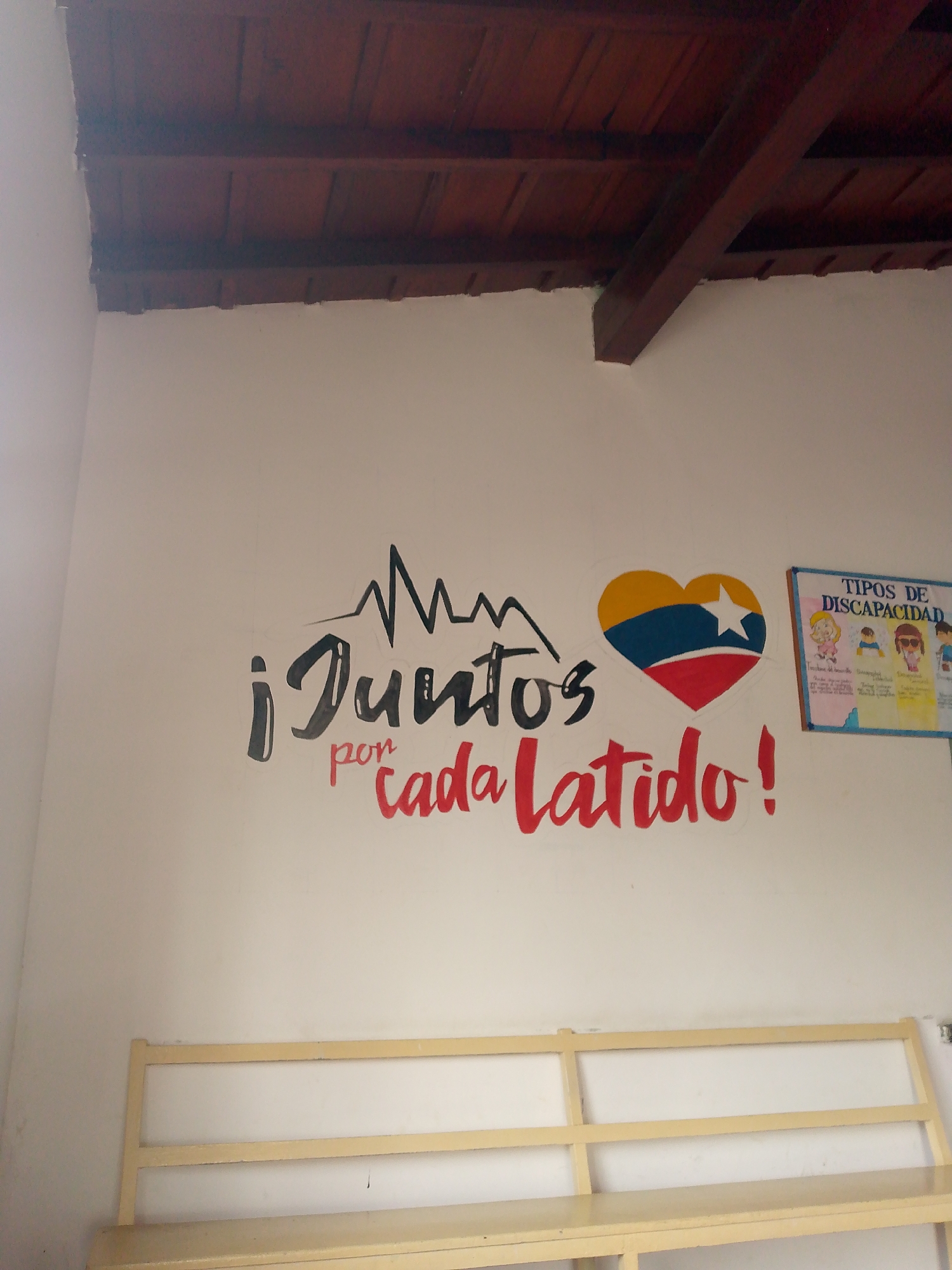 The image depicts an interior view of a building in Santa Ana, Venezuela. The walls are white, with a wooden ceiling. On the wall, there is a painted mural with the phrase "¡Juntos por cada latido!" The phrase translates to "Together for every heartbeat!" In the mural, there is a heart with the colors of the Venezuelan flag and a white star. Next to the mural is a poster titled "Tipos de Discapacidad," which translates to "Types of Disability." Beneath the mural, a light-colored wooden shelf is present. There are no people visible in the scene.