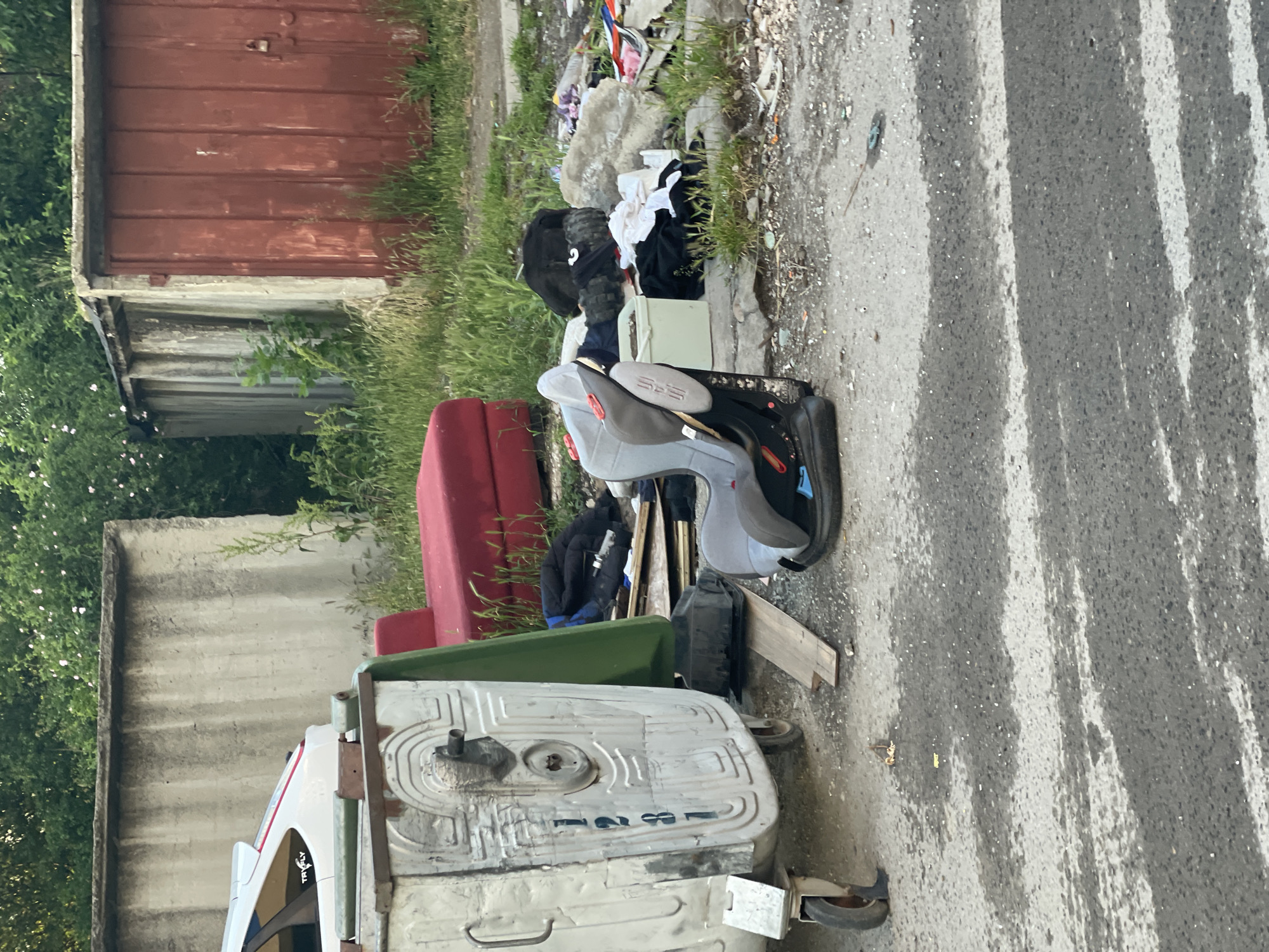 The image depicts a scene of urban decay in Sofia, Bulgaria. A damaged metal garbage container sits on a street, partially obscuring a red couch and a green bin. Discarded clothing and other trash are piled next to the garbage container. A child's car seat also sits among the debris. In the background are dilapidated structures, including a red garage and a concrete wall, overgrown with vegetation. The overall environment suggests neglect and disrepair. The street surface is worn and marked with stripes.