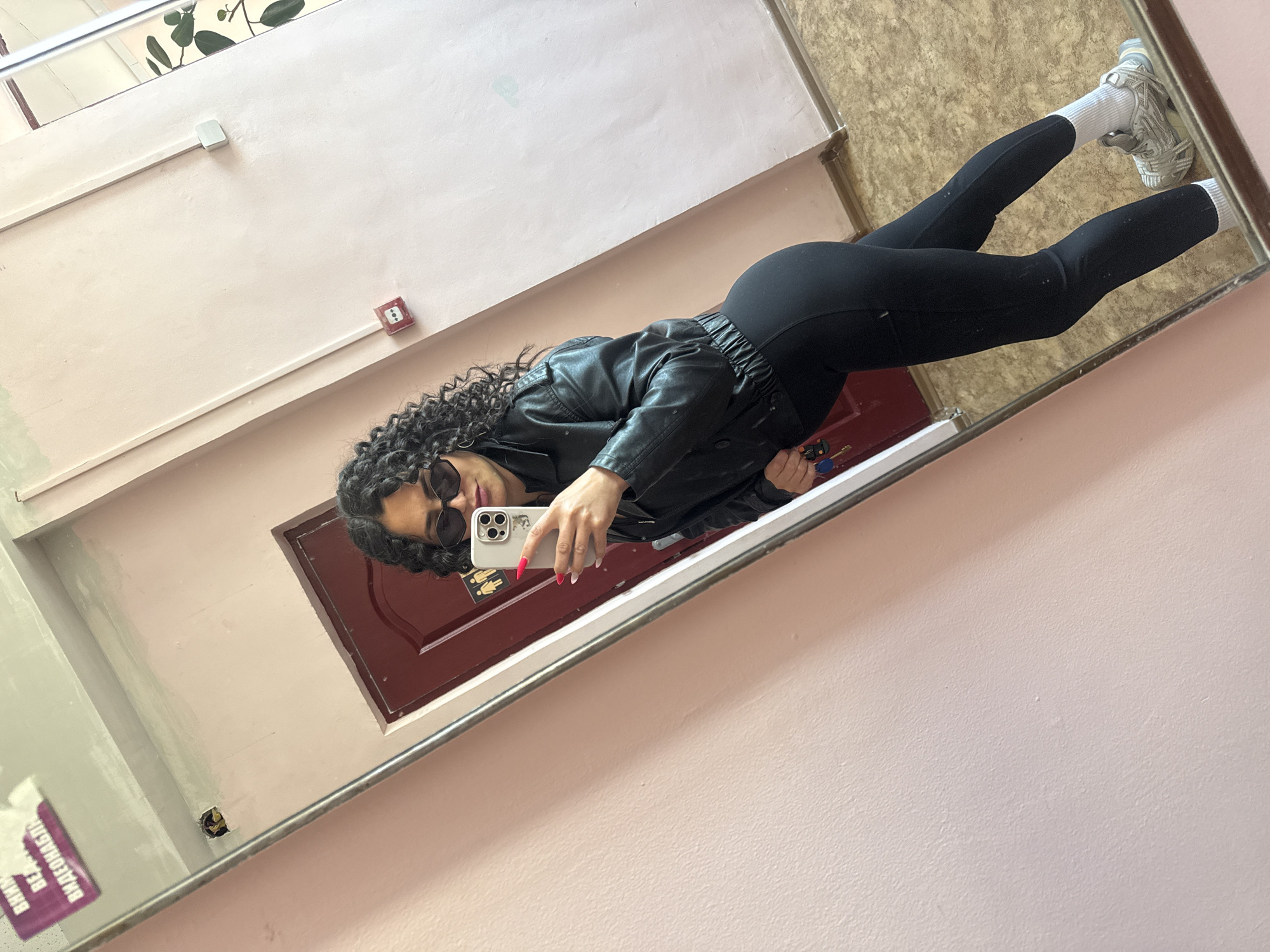 A young person is captured in a full-body mirror selfie, appearing to be in an indoor corridor or hallway in Chaikovsky, Russia. The image is tilted counter-clockwise, suggesting the person was holding the phone at an angle or the image was rotated after capture.
The subject has dark, curly hair and is wearing dark sunglasses. They are dressed in a black faux leather jacket, black leggings or athletic pants, white athletic socks, and chunky grey and white sneakers. They hold a light-colored smartphone with a case, revealing long fingernails painted in red and white. Their body is angled, and one leg is slightly bent at the knee.
The setting, reflected in the mirror, shows light pink or beige textured walls with exposed electrical conduit running across them. A white rectangular junction box and a red square emergency button or fire alarm are mounted on the wall. A dark brown or maroon door with paneling is visible behind the person's reflection. Above the door, a small sign with what appears to be gender symbols is partially visible. The floor is covered with a textured, light brown material, possibly linoleum. In the far upper left corner of the reflection, green leaves from a plant are visible, suggesting nearby greenery or a window.
On the far left side of the mirror, a purple sticker or poster is reflected, displaying Cyrillic text. Parts of it are legible, forming the phrase "ВНИМАНИЕ ОПАСНО!" which translates to "ATTENTION! DANGER!". This text strongly supports the provided location context of Russia. The overall scene suggests a casual moment captured in a public or semi-public indoor space.