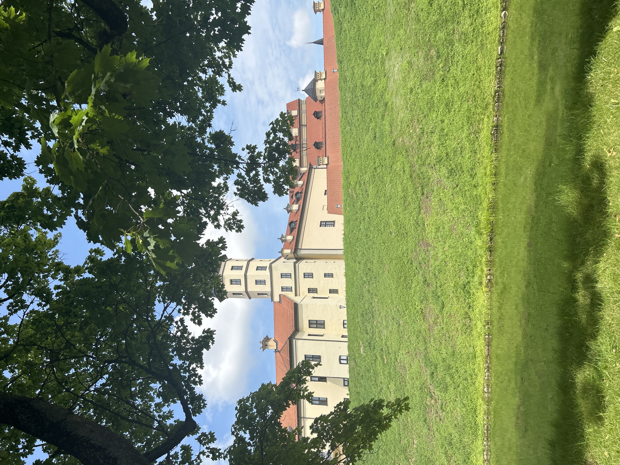 The image displays the Nesvizh Castle, a UNESCO World Heritage site in Nesvizh, Belarus. The castle, characterized by its pale yellow facade and red-tiled roofs, rises from a grassy incline. A prominent tower, part of the castle's architecture, stands tall. Lush green trees with dense foliage frame the top of the image, casting shadows. Below the incline, a tranquil body of water reflects the surrounding greenery and the sky. The sky is a clear blue, dotted with fluffy white clouds, suggesting a pleasant, sunny day. The overall scene conveys a sense of historical grandeur and natural beauty.