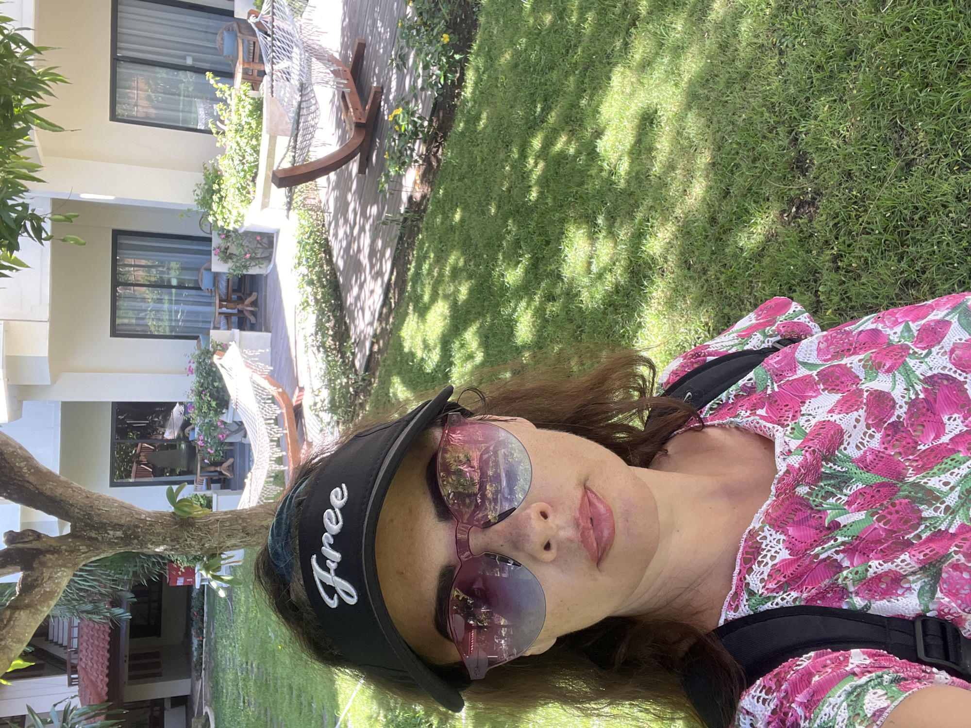 A woman takes a selfie outdoors, wearing a black visor with the word "Free" written in white script, large pink-tinted sunglasses, and a pink and white floral print top. She is standing in a grassy area with dappled sunlight. In the background, there is a building with balconies, hammocks, and outdoor furniture. The scene appears to be in a resort or hotel in Kuta Selatan, Indonesia.