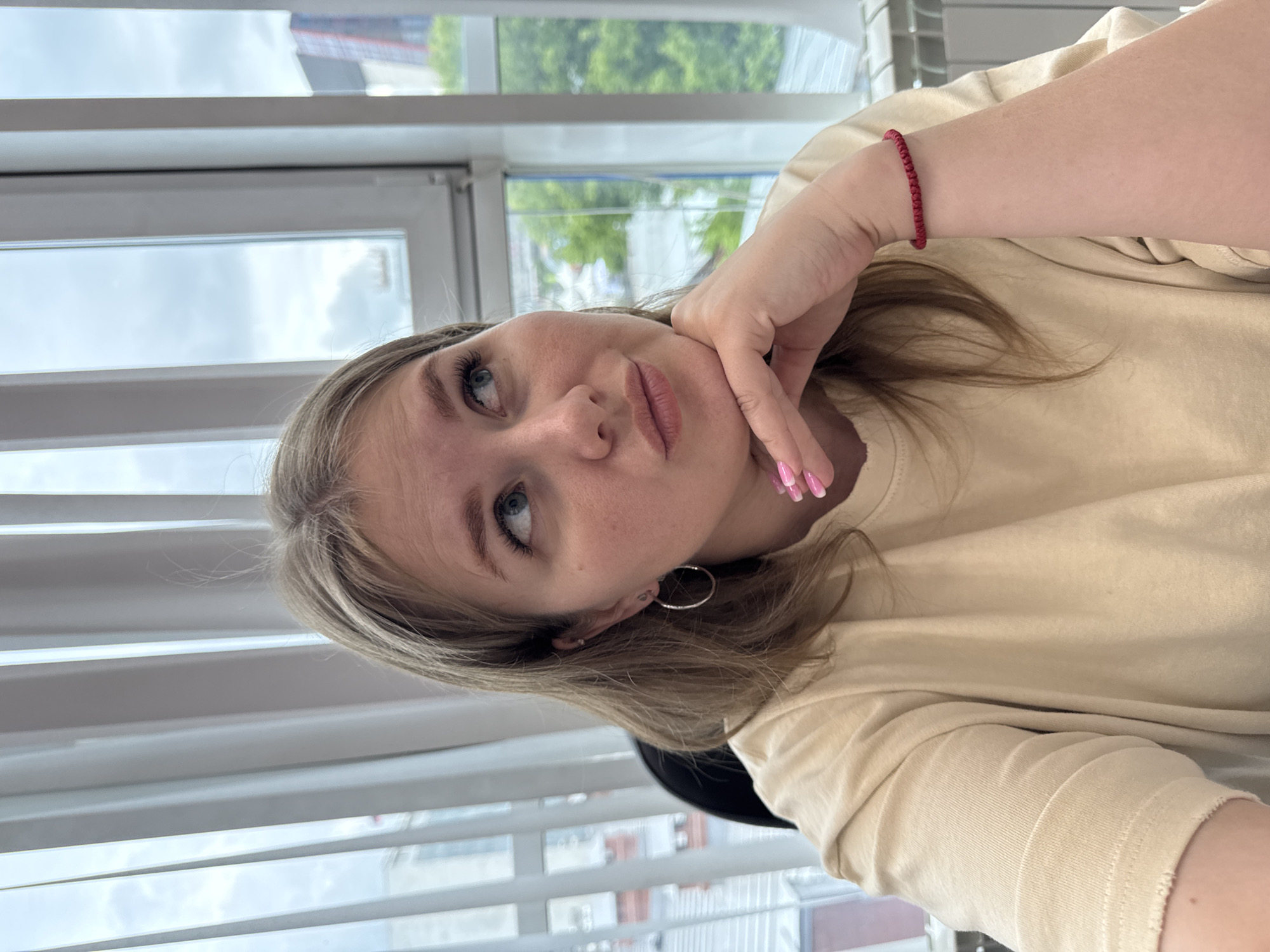 A young woman is posing for a selfie. She is fair-skinned with blond hair and blue eyes. Her left hand is supporting her face and she is wearing a red bracelet on her left wrist. She has silver hoop earrings in her ears. She is wearing a beige long-sleeved shirt. The location is a room with venetian blinds covering a window. Outside the window, foliage is visible. The location is likely in the city of Perm, Russia based on prior knowledge.