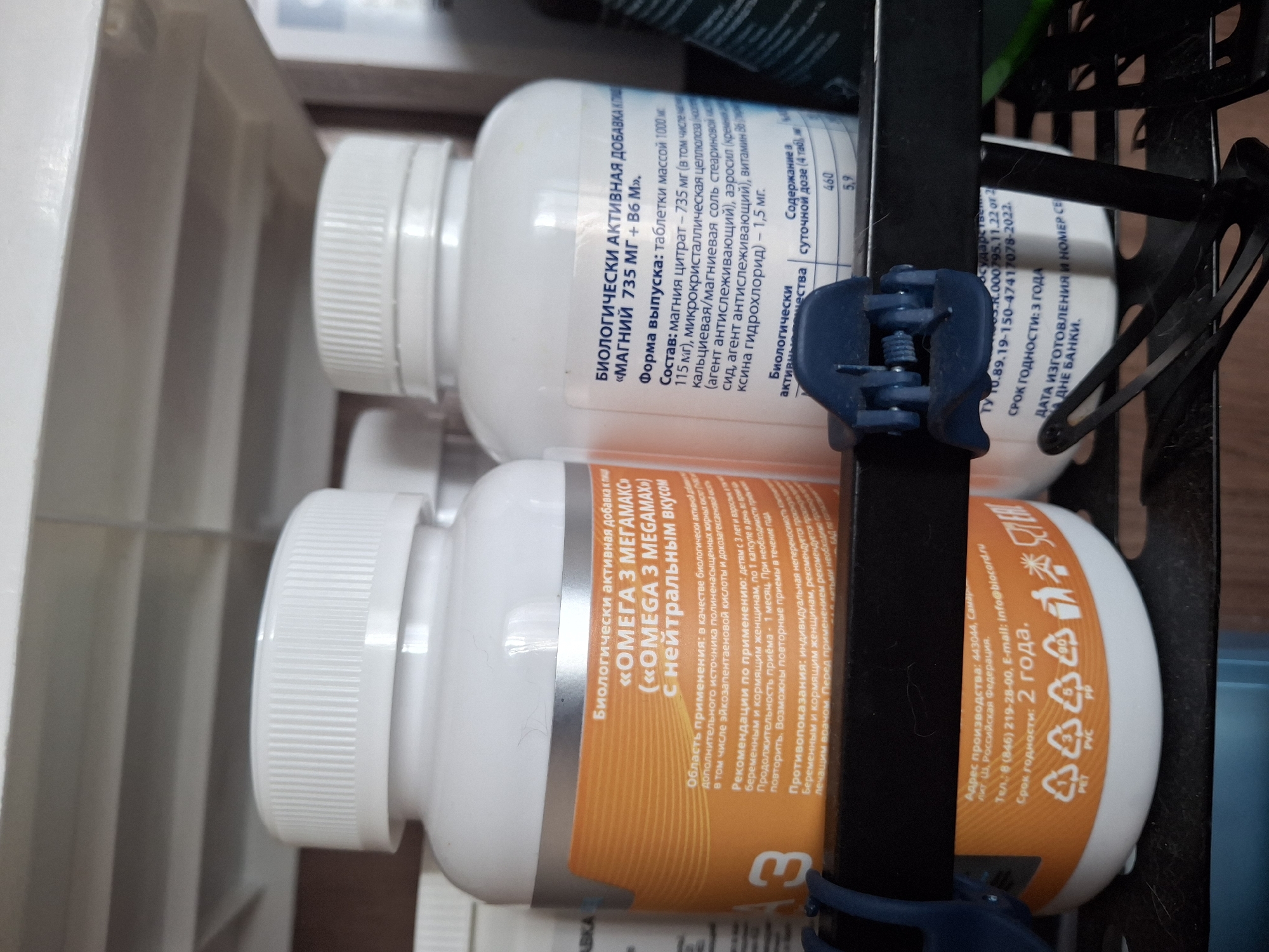 The image shows two white plastic bottles containing dietary supplements, placed on a black metal shelf. The bottle on the left has an orange label with white text, partially visible. The text on this label reads "ОМЕГА 3 МЕГАМАКС" (Omega 3 Megamax) and "С НЕЙТРАЛЬНЫМ ВКУСОМ" (with neutral taste). It also contains information about the area of application, recommendations for use, and contraindications. Below this, the production address is listed as "443044, Самарская обл, г. Тольятти, Российская Федерация." (443044, Samara region, Tolyatti, Russian Federation). The expiration date is indicated as "2 года" (2 years). The bottle on the right has a white label with black text. The visible text on this label reads "БИОЛОГИЧЕСКИ АКТИВНАЯ ДОБАВКА К ПИЩЕ" (Biologically active food supplement) and "«МАГНИЙ 735 МГ + В6 М»" (Magnesium 735 mg + B6 M). It also includes information on the form of release, composition, and biological activity. A small blue clip is attached to the shelf in front of the bottles. The background consists of shelves, suggesting a retail or storage environment.