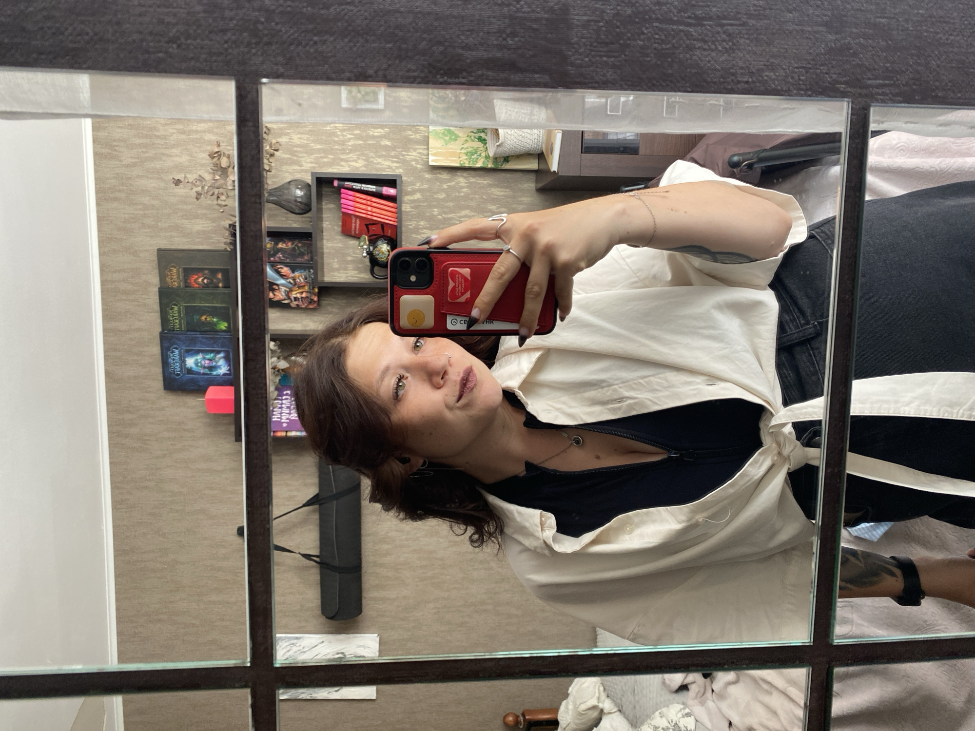 A woman with dark hair takes a mirror selfie in a room. She is wearing a black top and an open white button-down shirt tied at the waist. She is holding a red phone in her right hand, which has a phone case with a yellow sun sticker and a red heart sticker with a white outline. She is wearing silver rings on her right hand and a silver bracelet on her left wrist. She has a tattoo on her left arm. In the background, there are bookshelves with books, a red candle, a vase, and a window with curtains. The room appears to be a bedroom, with a bed visible on the left side of the image. The overall setting suggests a personal living space. The city of Chaikovsky, Russia, is mentioned as the location.