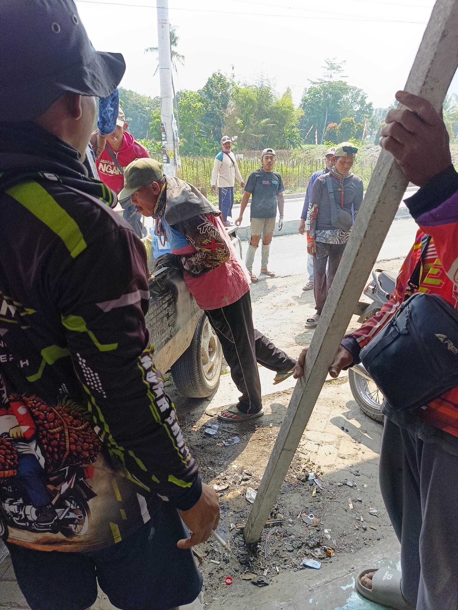 In Sukorini, Indonesia, a group of men are gathered outdoors on a sunny day, seemingly engaged in manual labor. The scene is populated by several individuals, some of whom are smoking. One man, wearing a red shirt with text that appears to read "SEDULUR NGA," is leaning against a dark-colored vehicle. Another man, in a pink vest and dark pants, is also near the vehicle and appears to be smoking. A third individual, wearing a black hat and a patterned long-sleeved shirt, is in the foreground, facing away from the camera. A thick, light-colored plank of wood is being held vertically by at least two people, one on the right side of the frame and another, whose face is obscured, on the left. The ground is covered with dirt, debris, and cigarette butts. In the background, there are trees, a utility pole, and what looks like construction work in progress. The overall impression is a candid snapshot of a work scene, possibly involving construction or road maintenance, with a casual atmosphere among the workers.